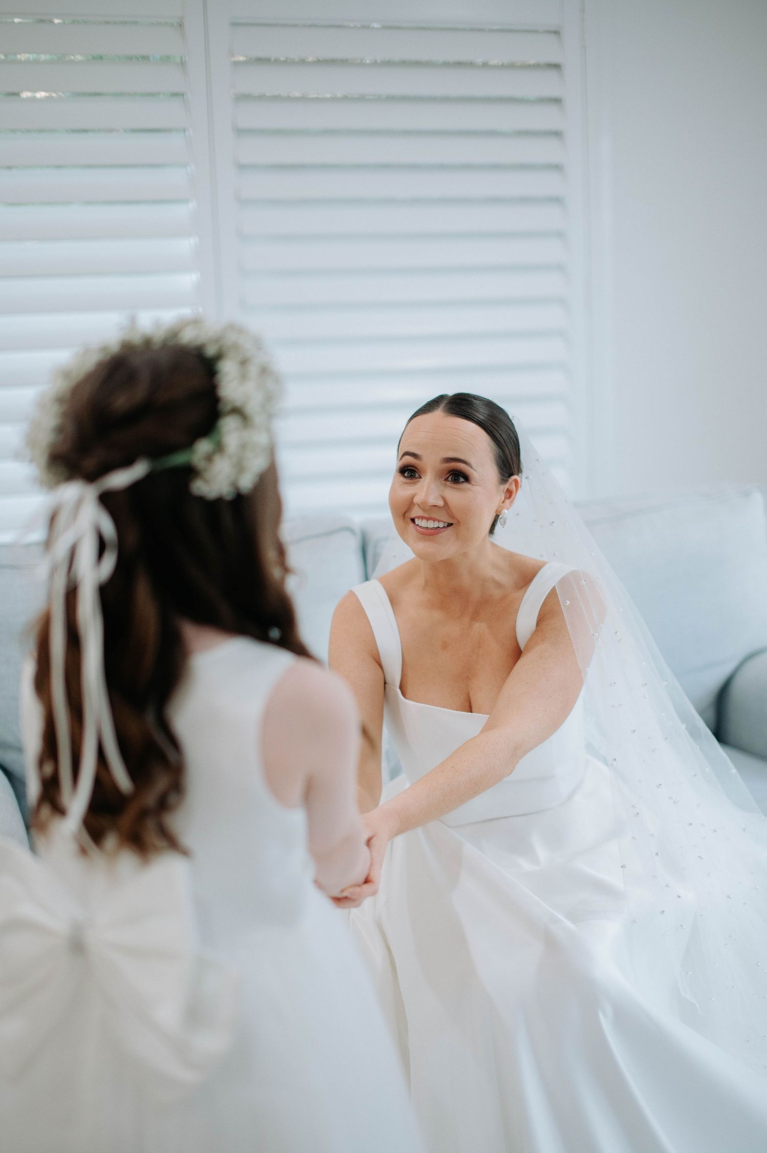 A tender moment between two figures in white with one wearing a bridal veil and flower crown in a light-filled room.