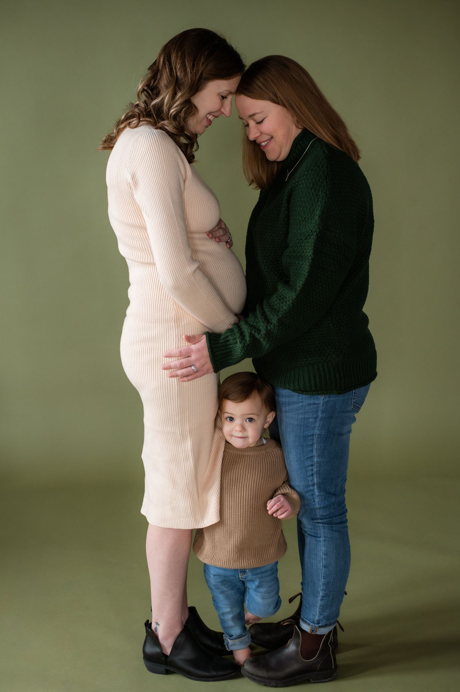A heartwarming family moment against a green backdrop as two parents share an intimate moment while their toddler looks on.