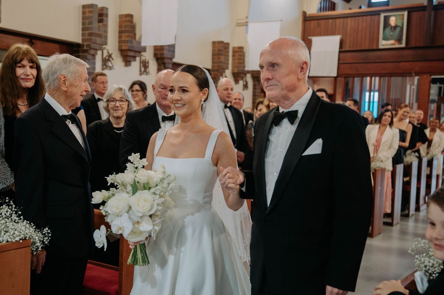 A bride in a white wedding dress walks down the church aisle holding a bouquet of white flowers.
