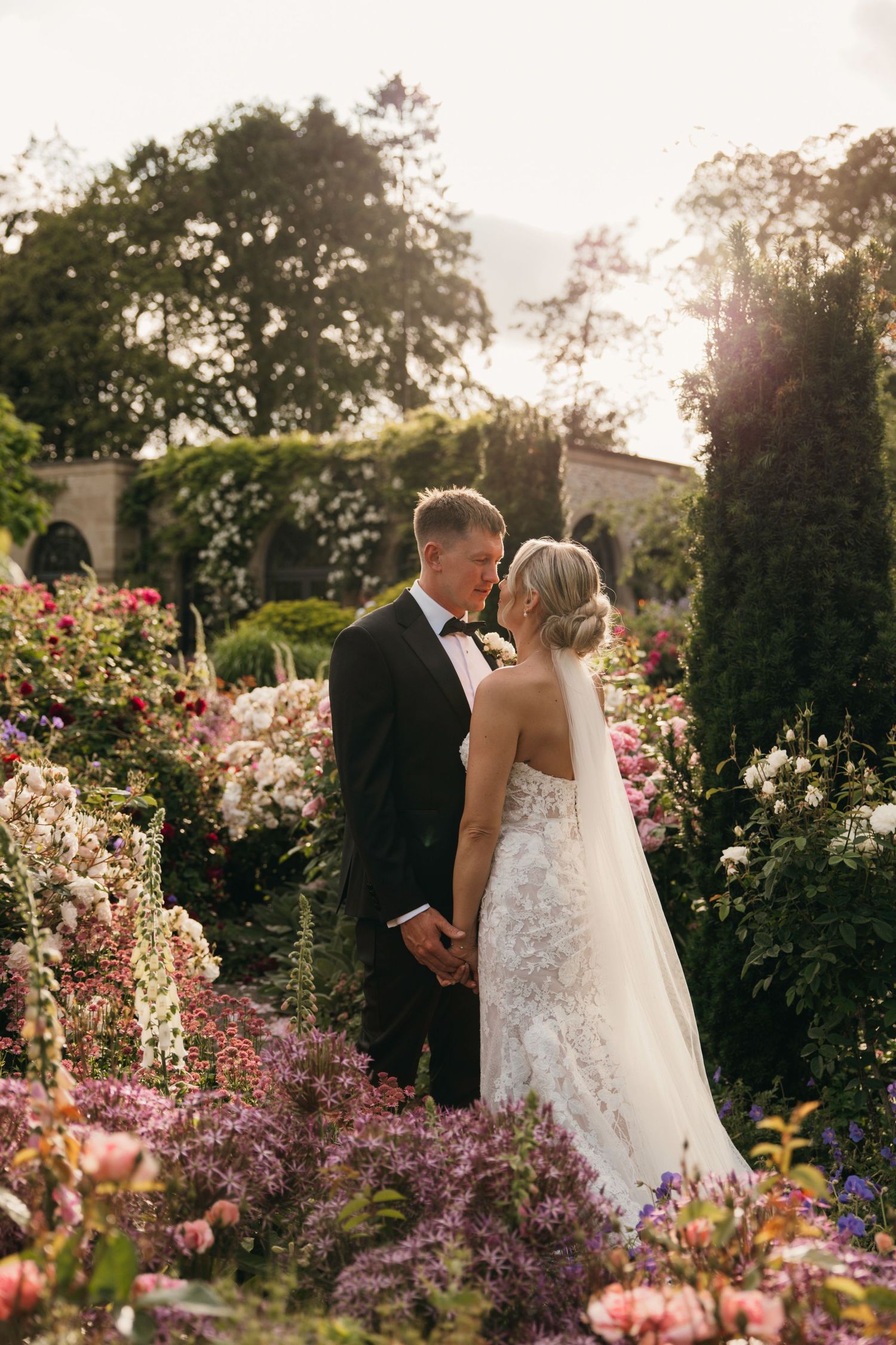 Bride and groom holding hands among colorful garden blooms at golden hour, bride in lace gown with veil.