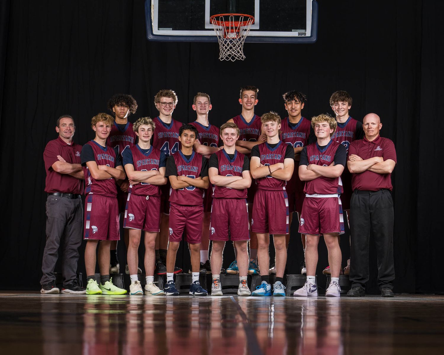 A basketball team wearing maroon uniforms poses for a group photo on a hardwood court in front of a backboard.