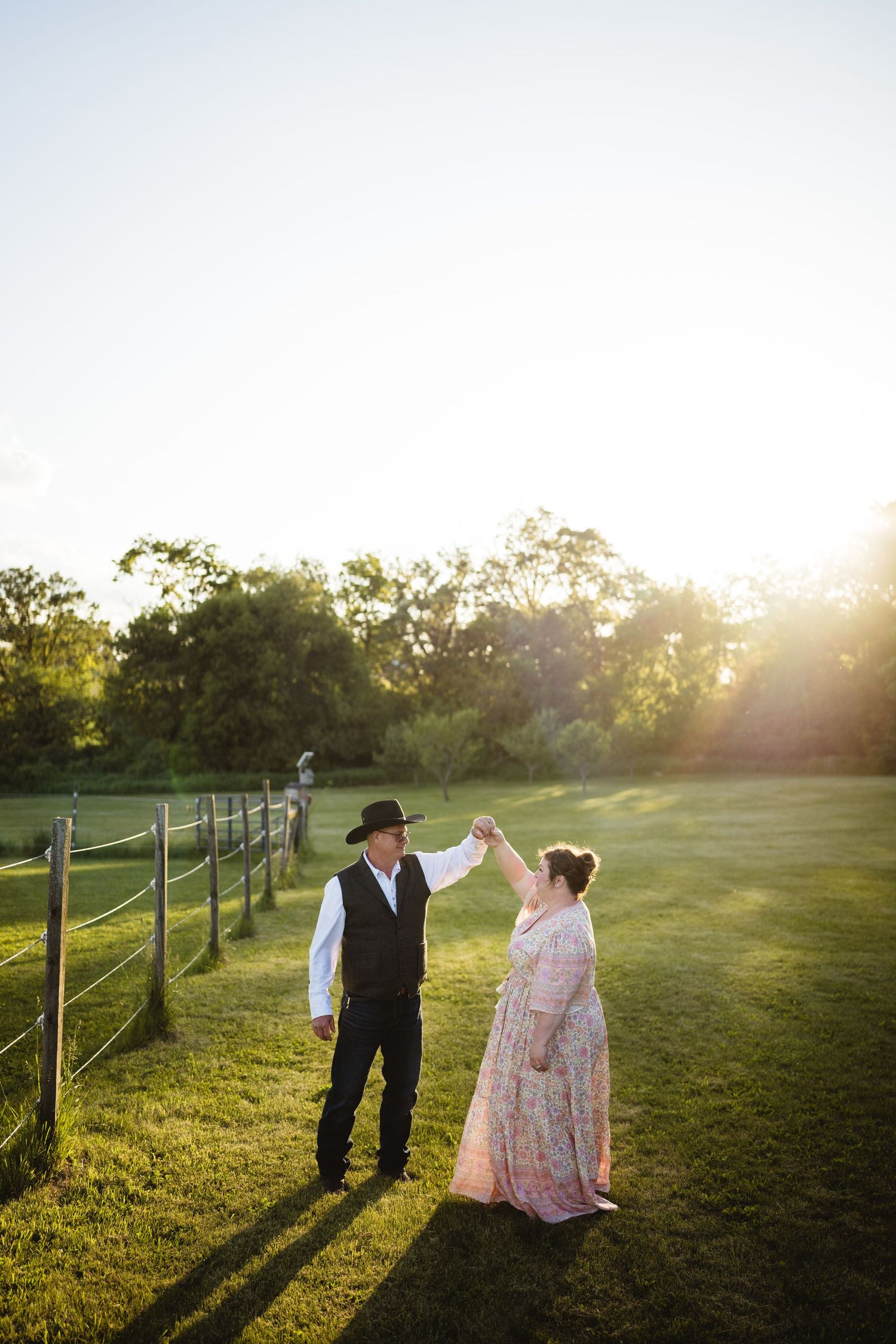 A couple in period dress dances together at sunset in a pastoral setting with wooden fencing and trees in the background.
