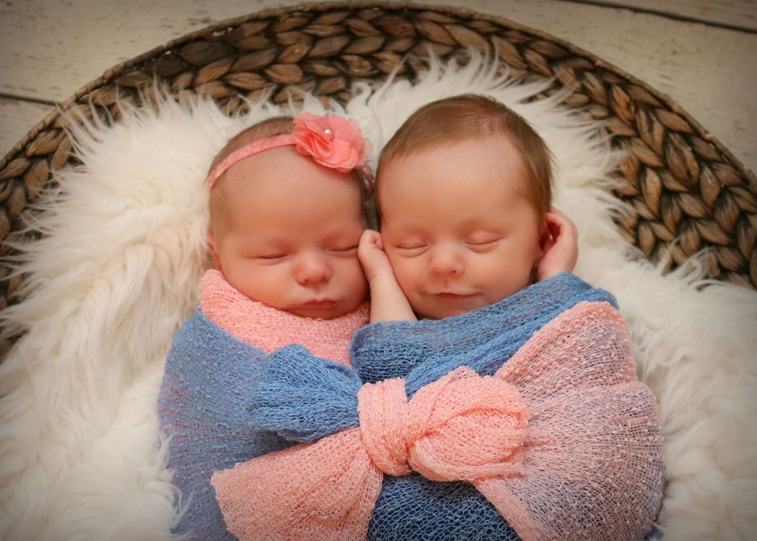 Twin newborns sleeping peacefully wrapped in pink and blue swaddles on a round woven mat.