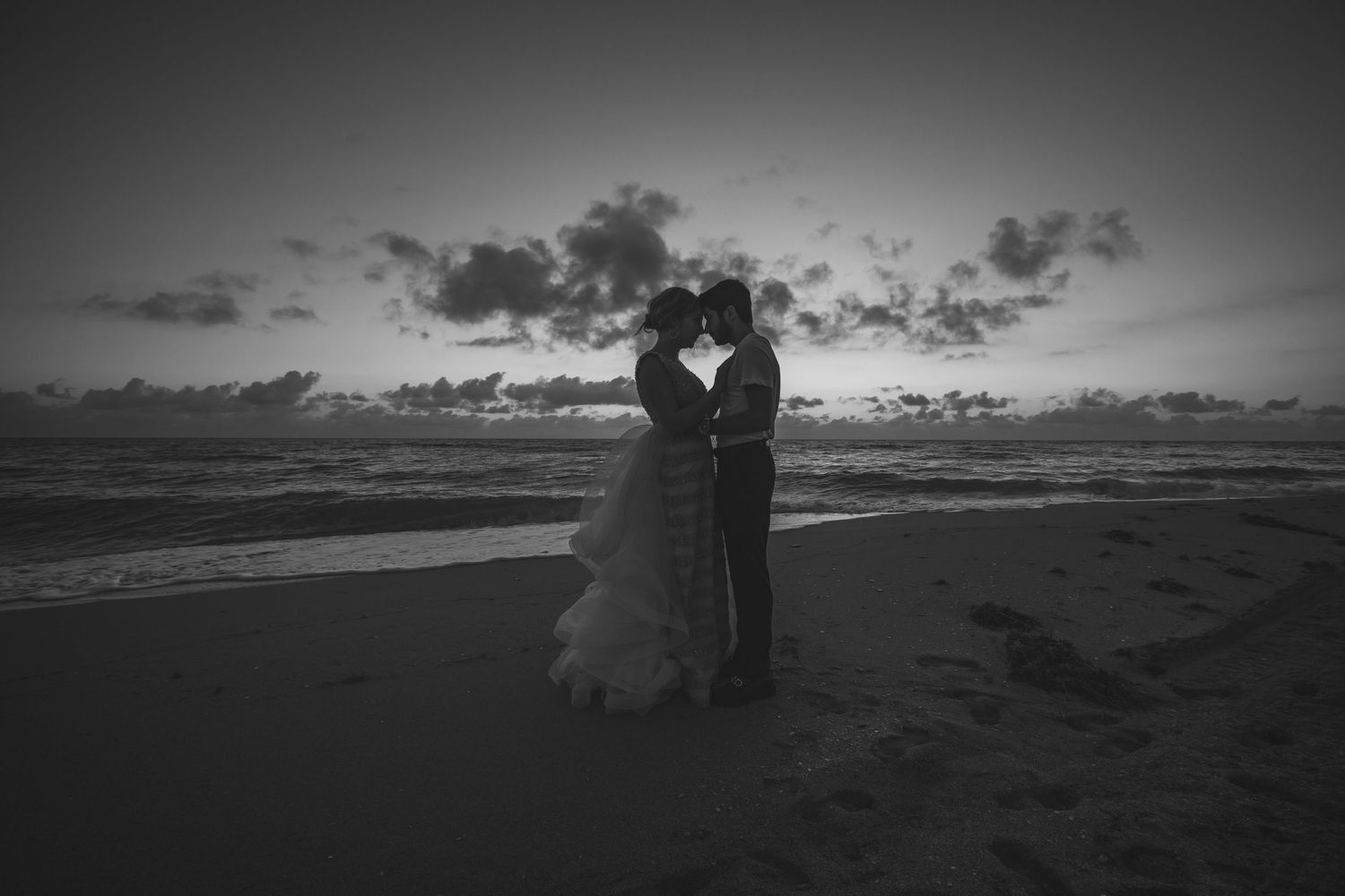 A silhouetted couple shares an intimate moment at sunset on a beach with dramatic clouds in black and white.