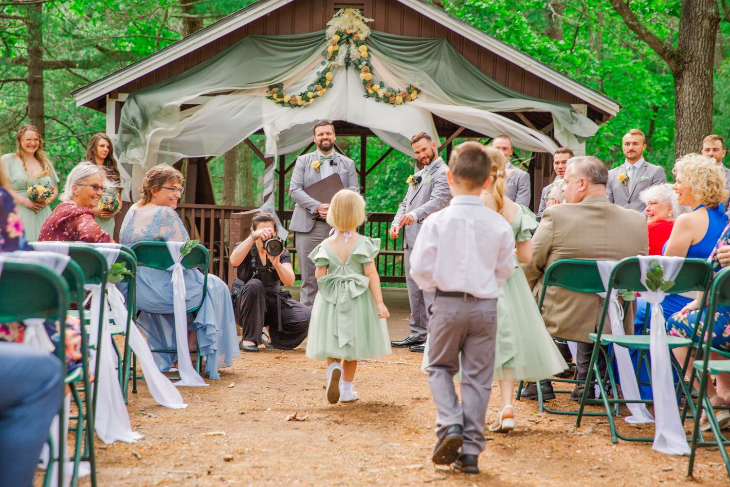 A rustic outdoor wedding ceremony in a wooded setting with guests gathered around a decorated gazebo in the forest.