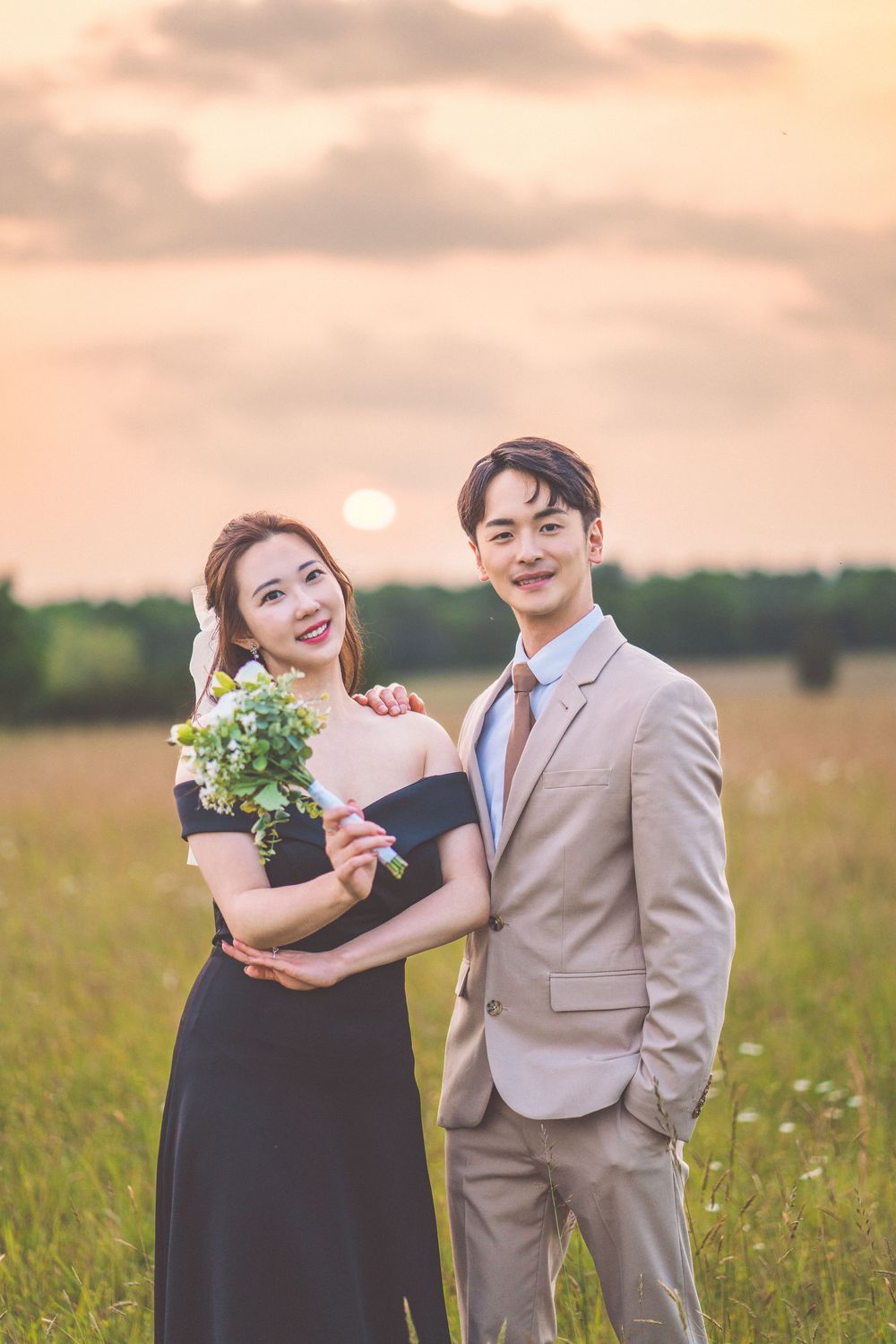 Couple in formal wear posing together in a sunlit meadow with a bouquet of flowers.