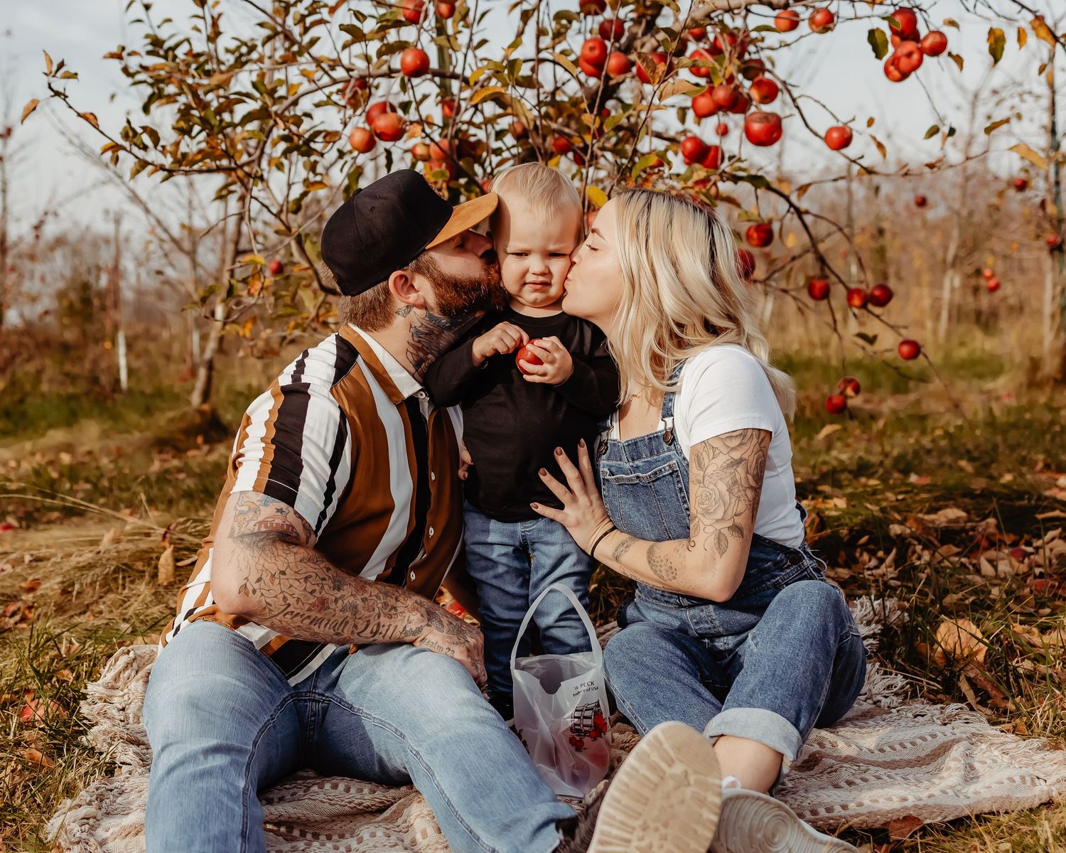 Family sits on blanket under red autumn berries sharing intimate moment in golden sunlit orchard.