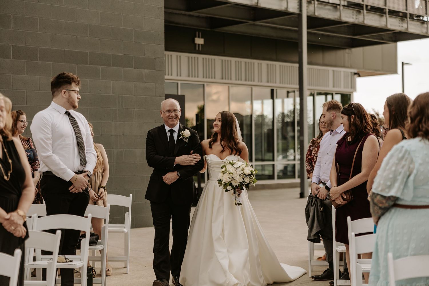 Wedding ceremony taking place outdoors in front of modern building with white chairs and gathered guests.