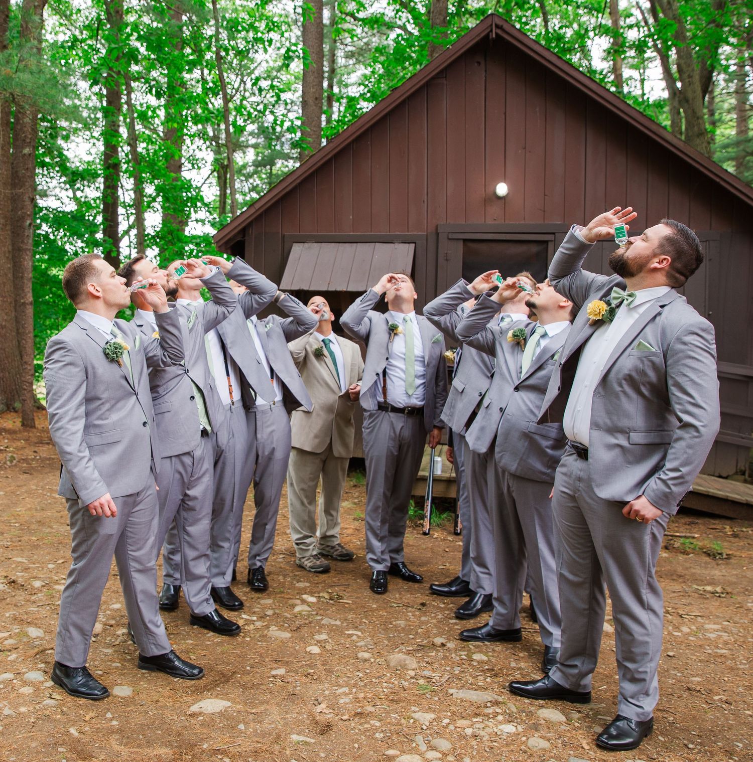 Group wearing matching light gray suits tilt their heads back while posing for a fun photo in front of a brown cabin.