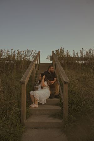 A couple sits intimately on wooden stairs surrounded by tall grass during golden hour.