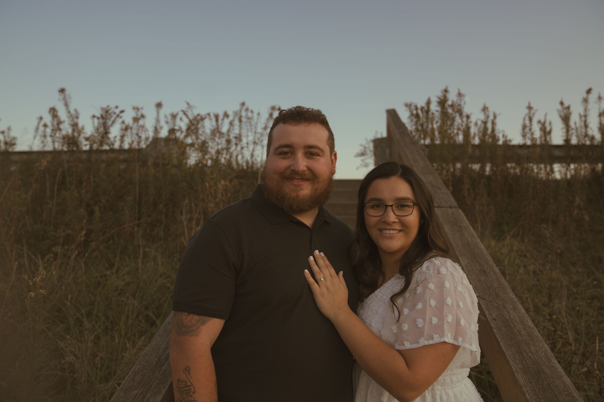 A couple stands close together on a wooden walkway during sunset.