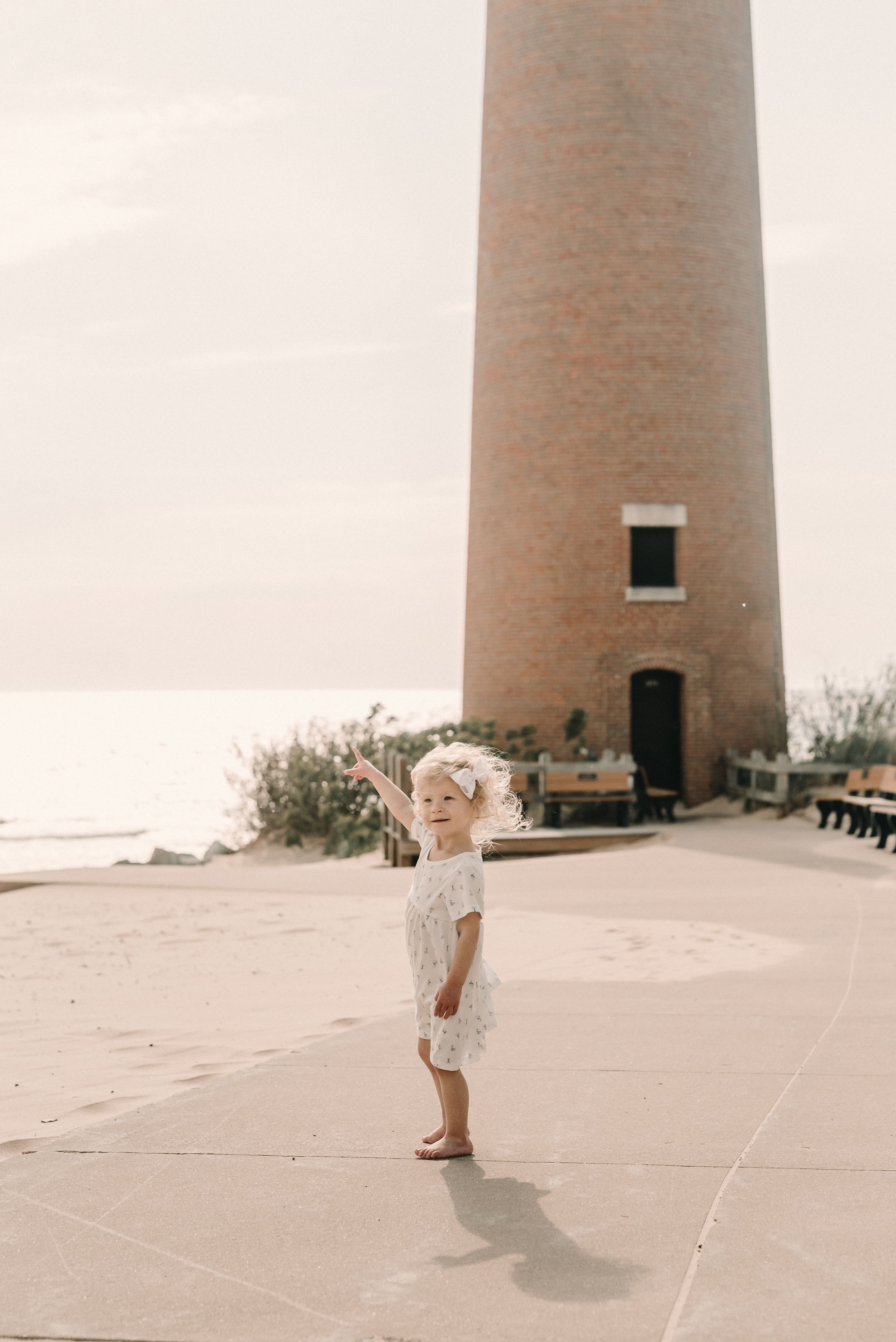 A toddler in a white dress stands alone in front of a tall lighthouse tower on a sunny afternoon.