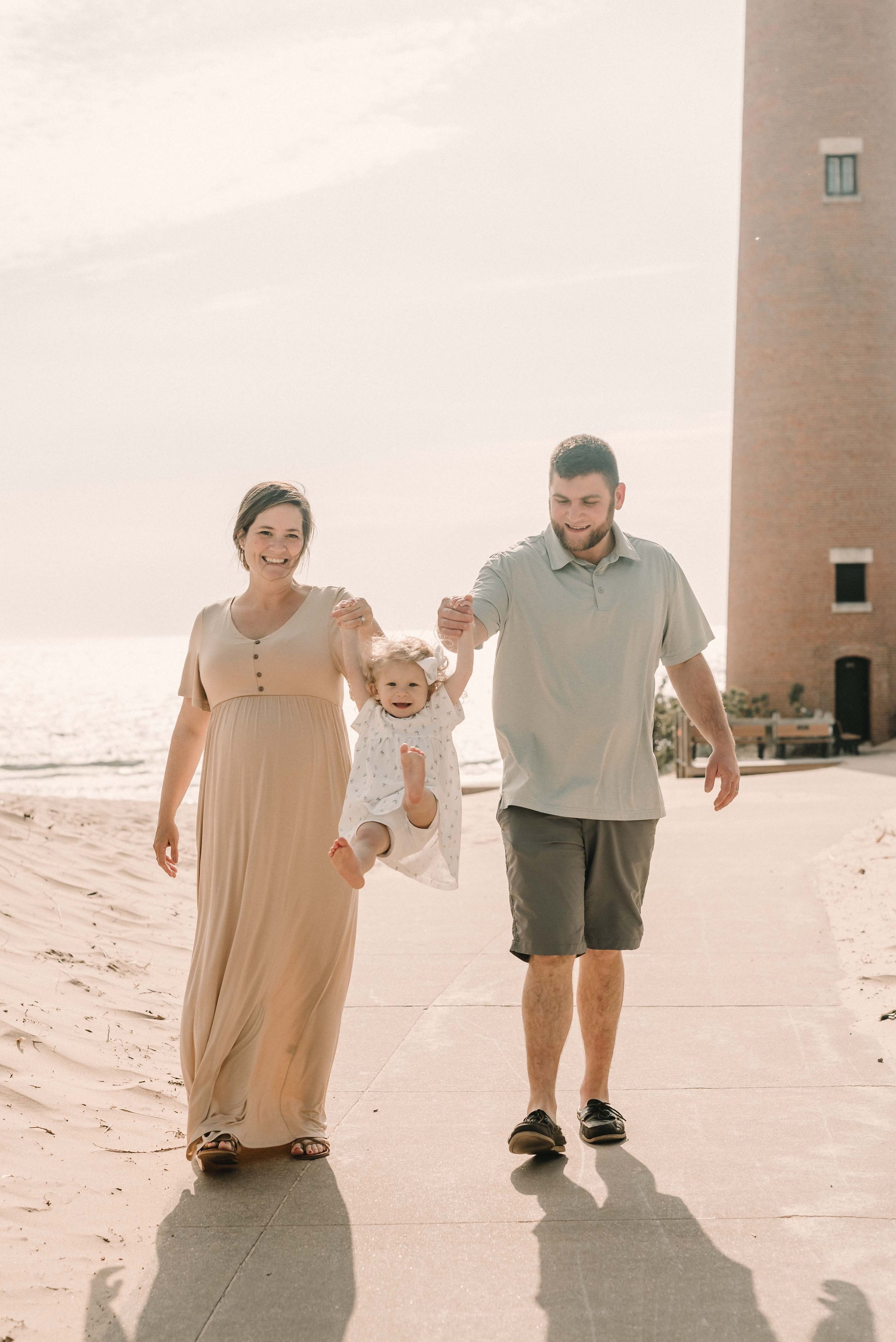 A family of three walks together on a sandy path near a brick lighthouse on a bright sunny day.