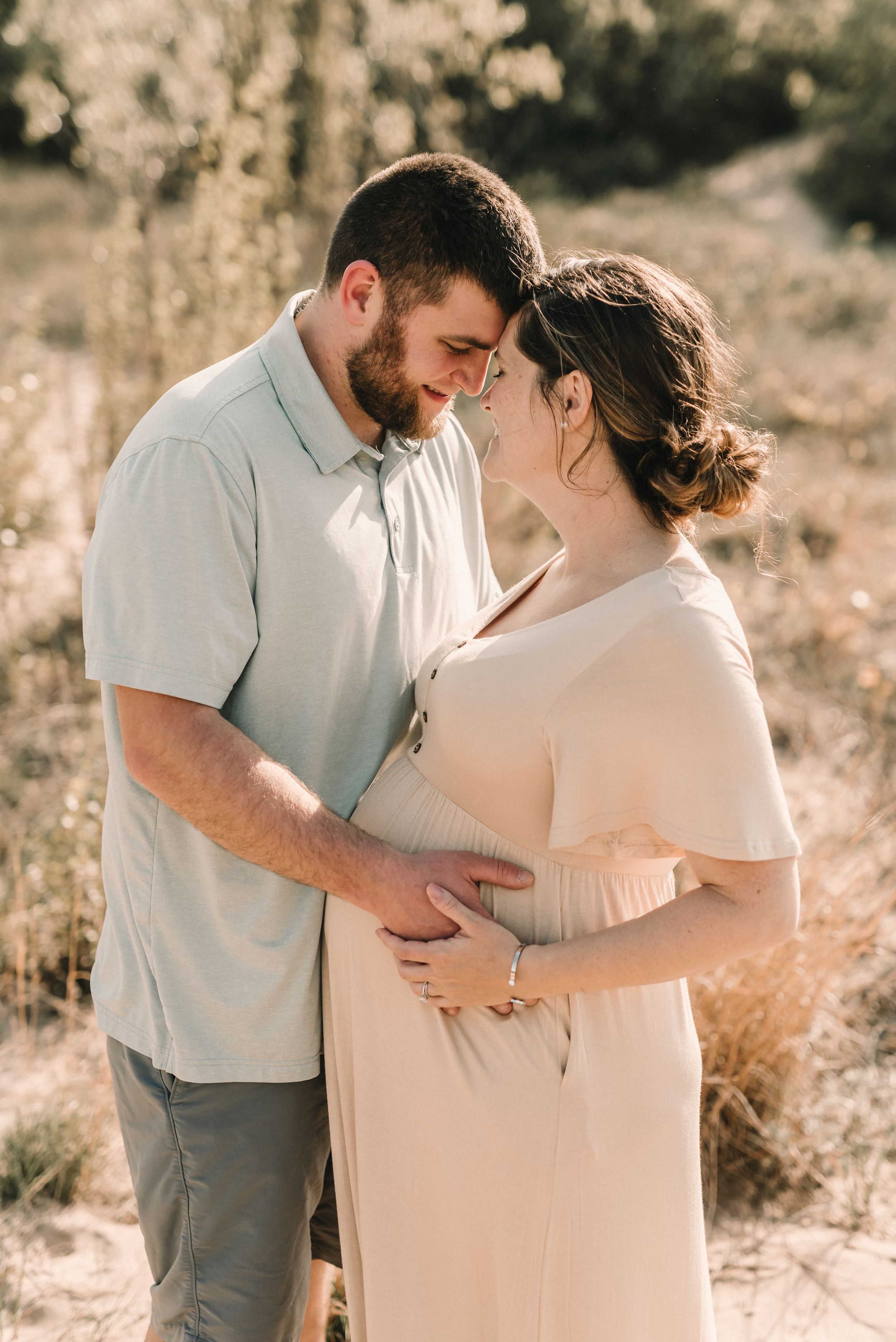 A couple shares an intimate moment outdoors in soft lighting, with the woman wearing a blush dress and cradling her belly.