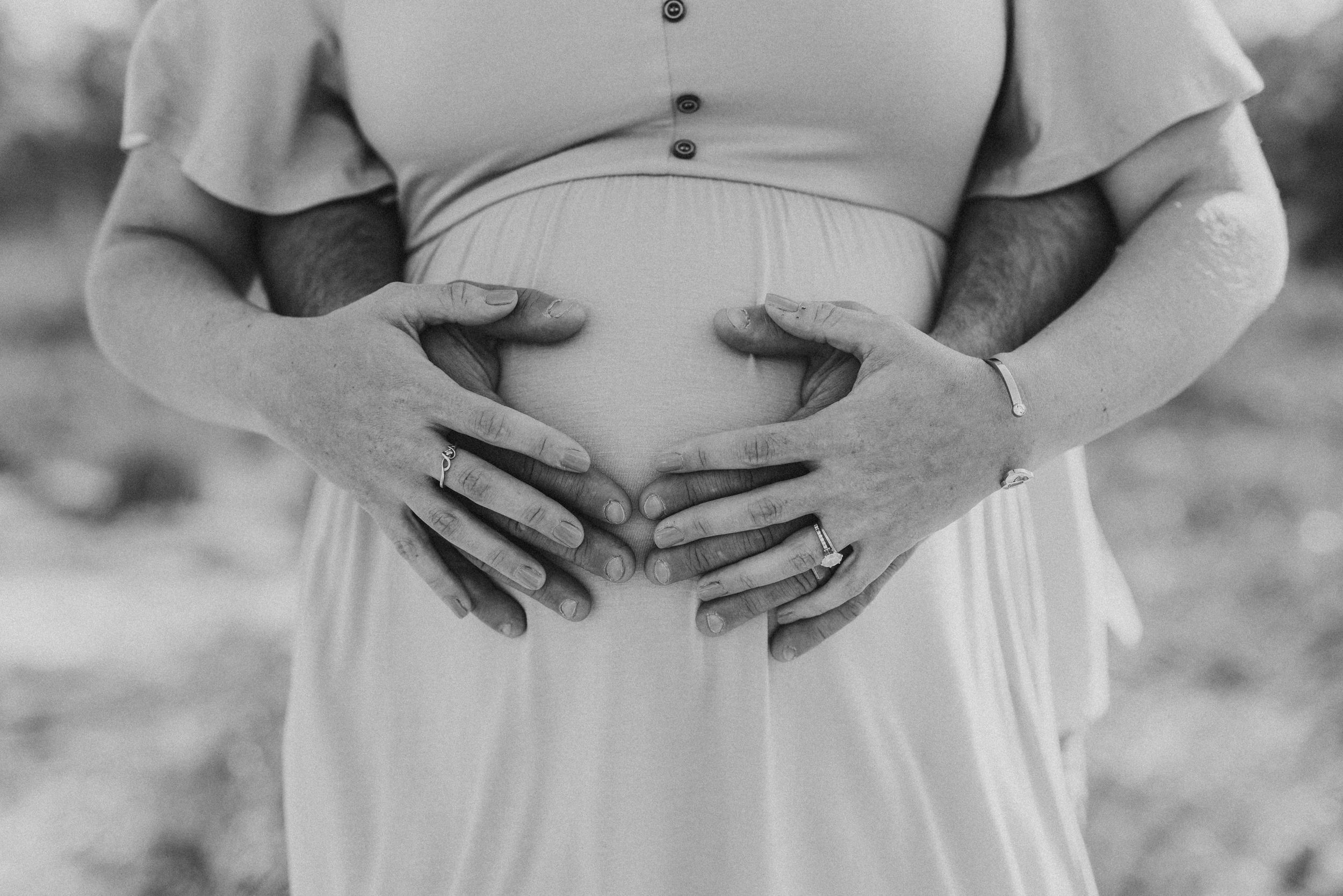 Black and white photo of hands gently cradling a pregnant belly in flowing dress outdoors.