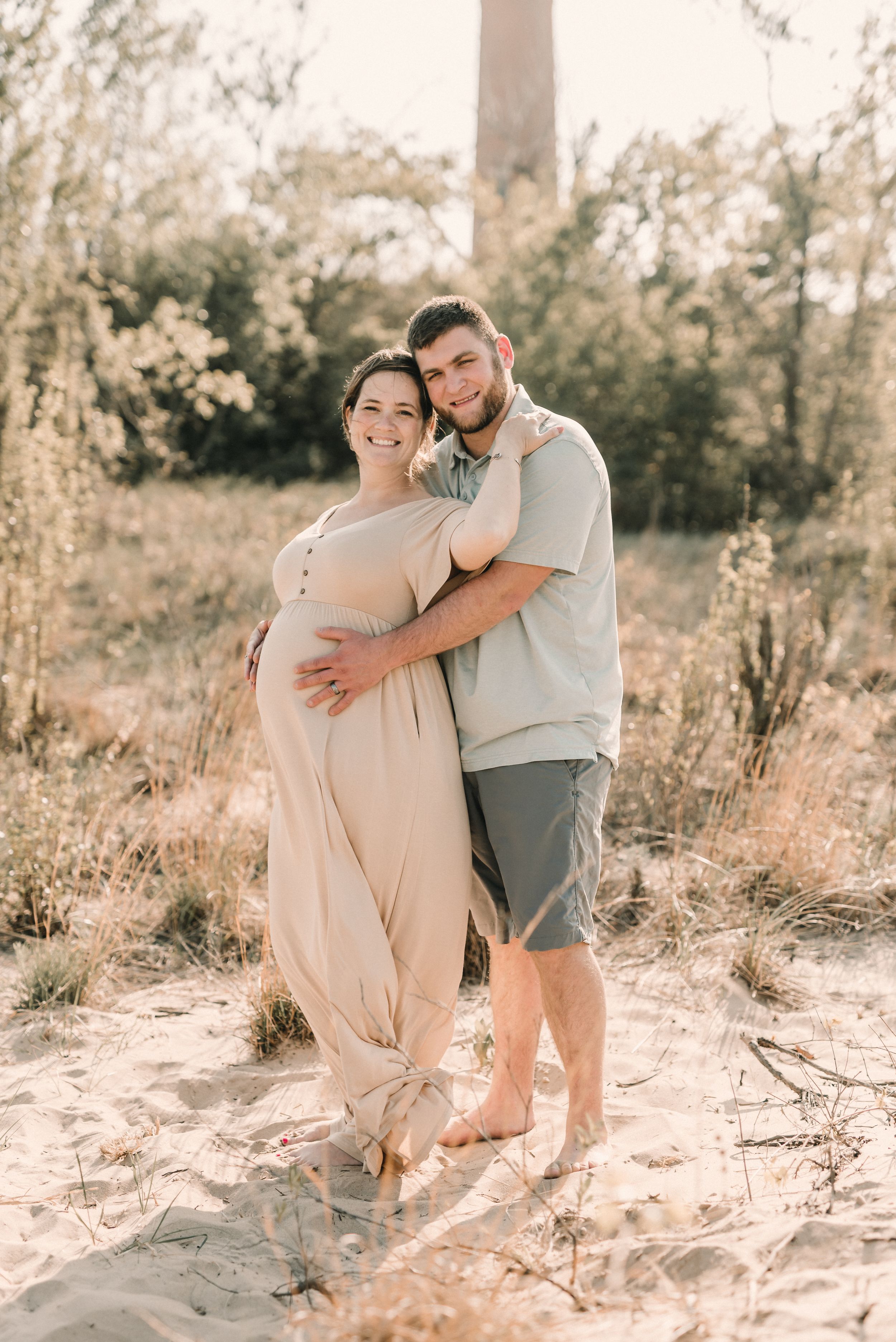A couple embraces on a sandy beach during a warm sunset maternity photoshoot.