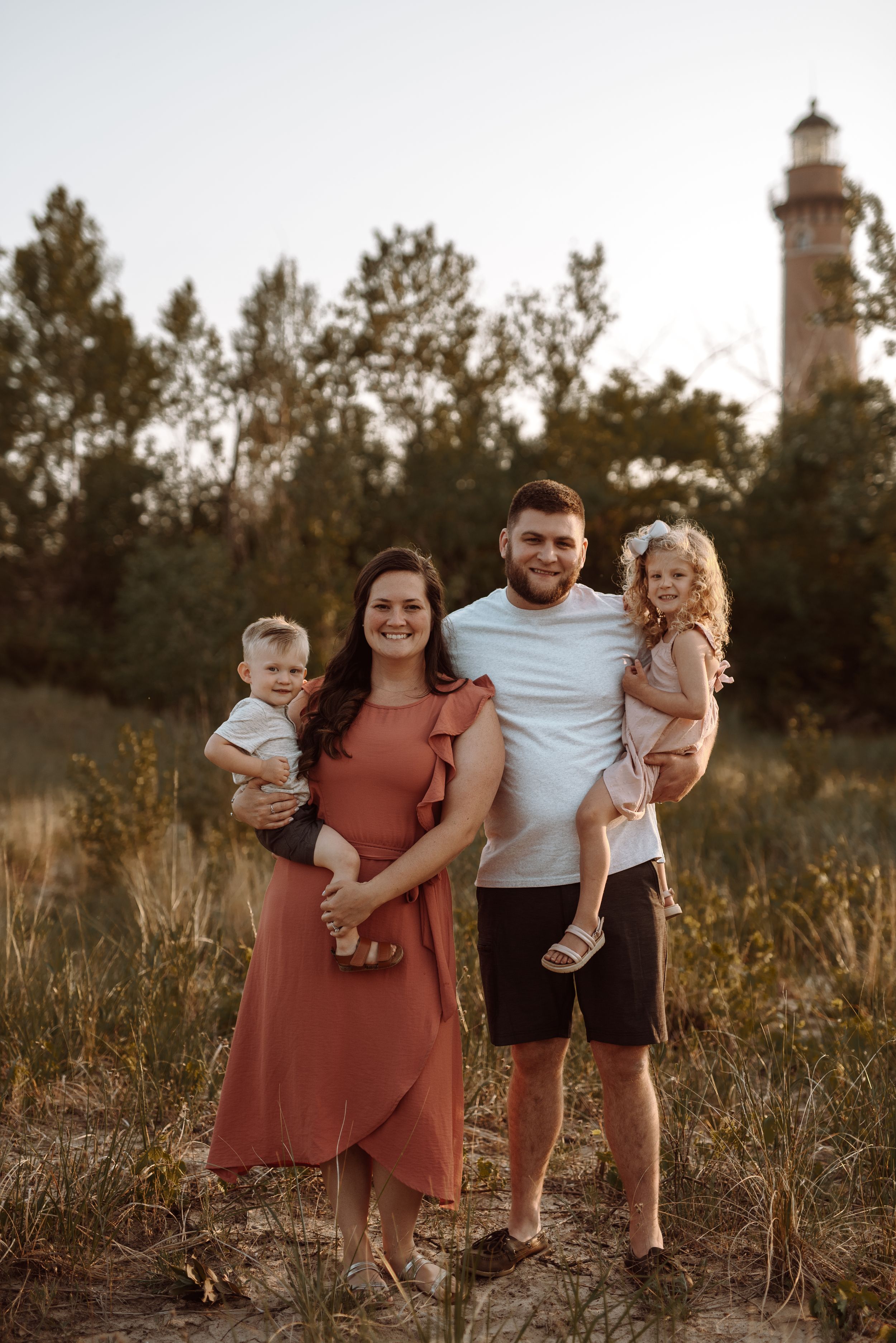 Family portrait session at sunset in a field with tall grass and a lighthouse in the background.