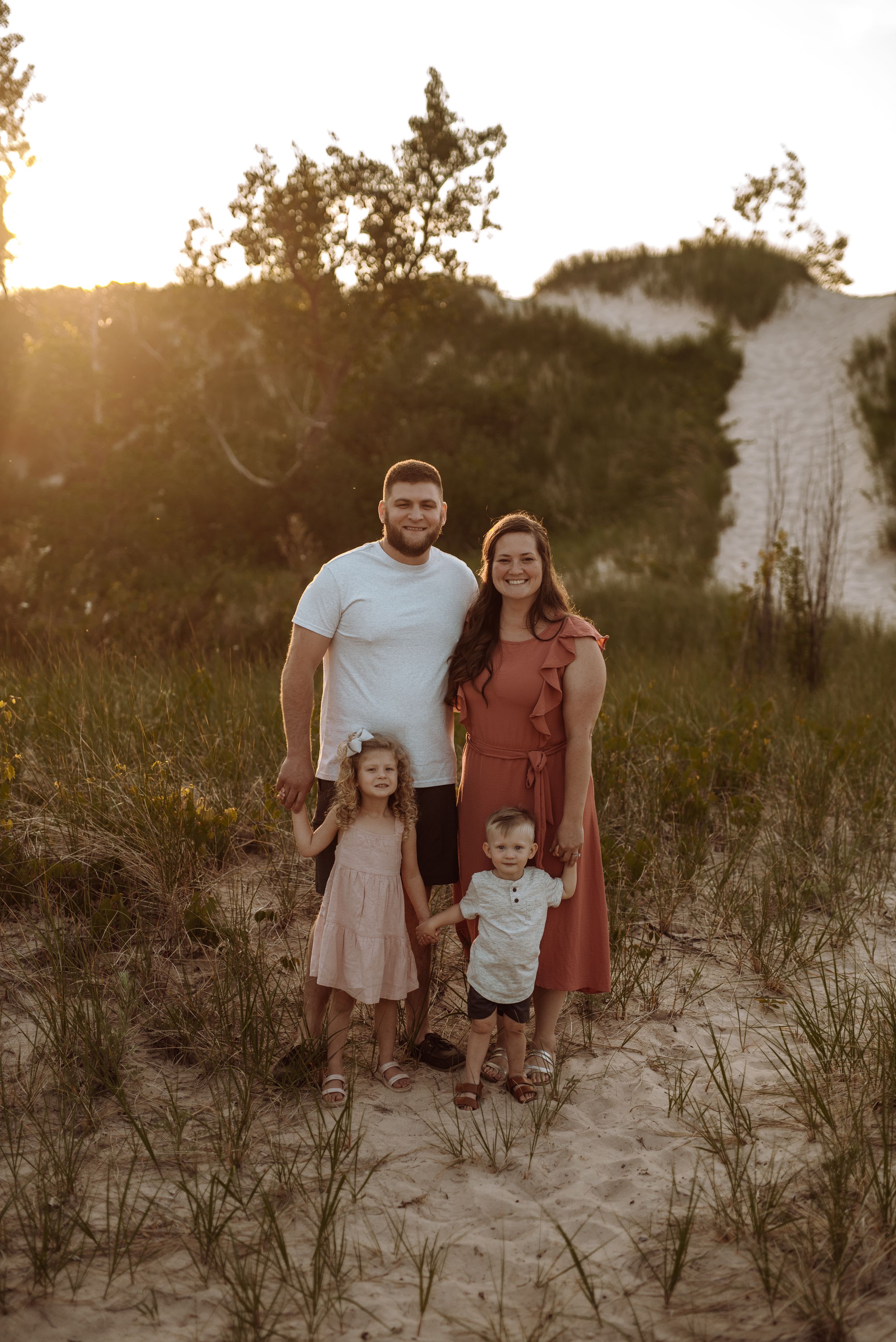 A family stands together on sandy dunes at sunset with trees silhouetted in the background.