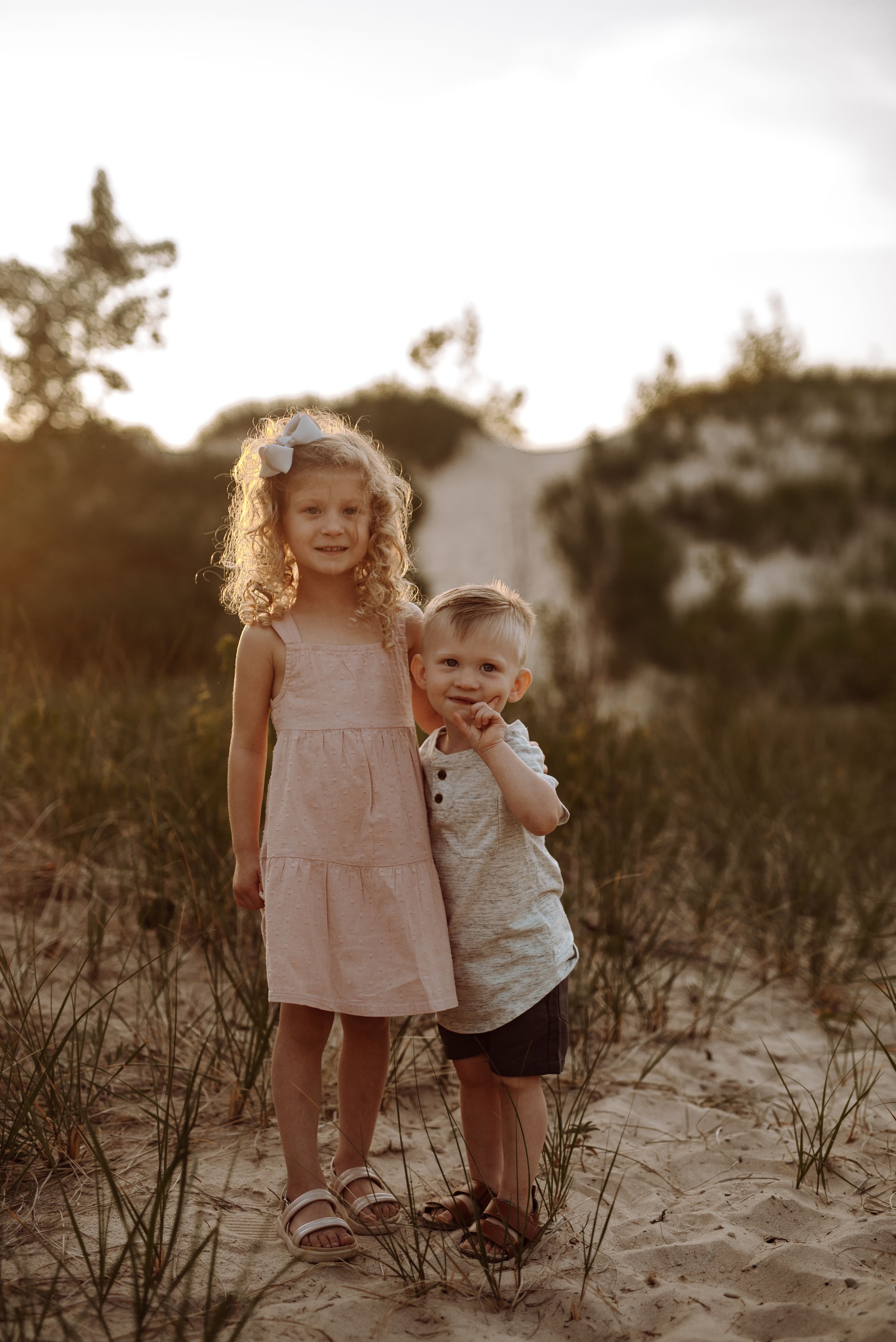 Two children standing together on sandy ground at sunset with trees in background creating warm golden atmosphere.
