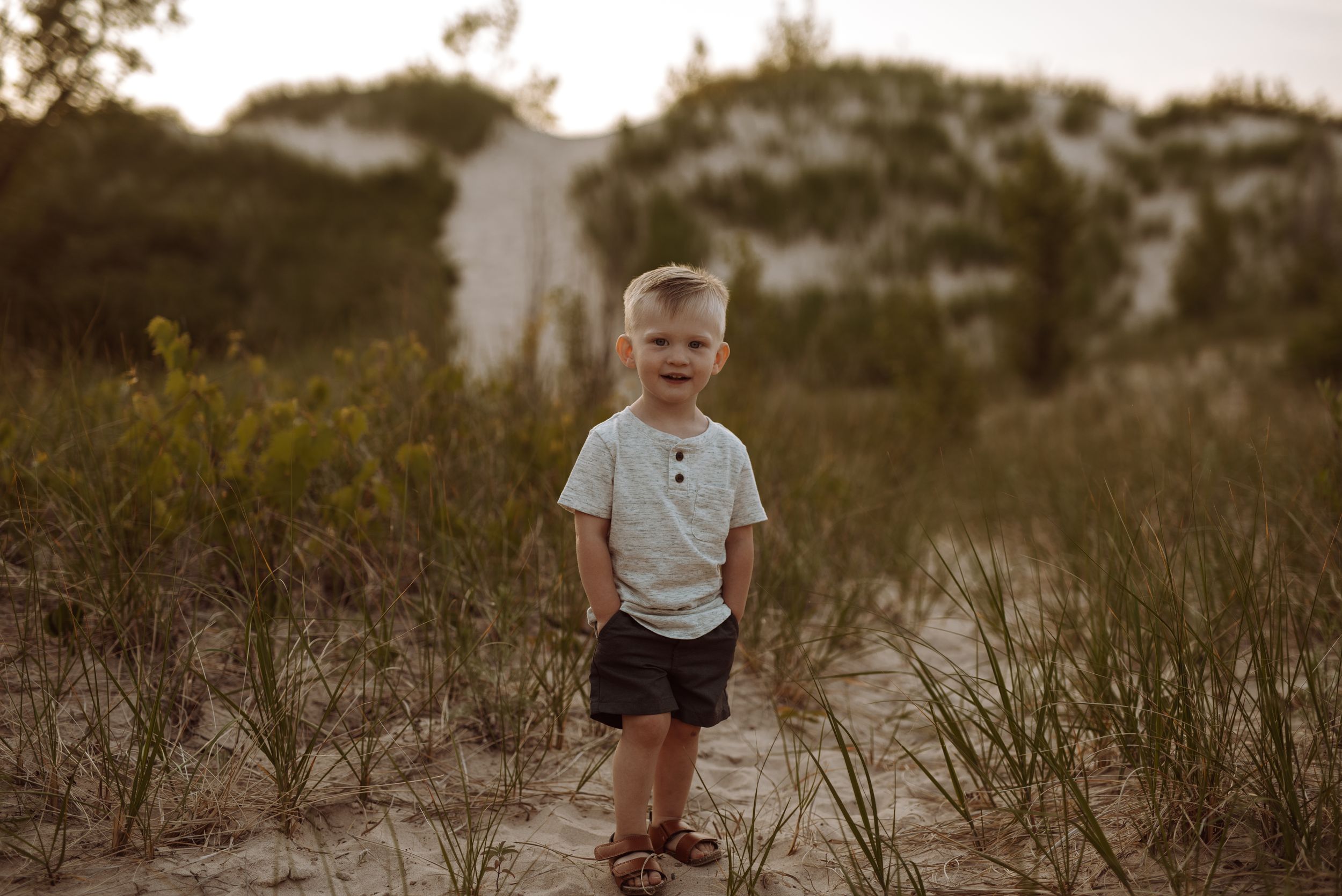 A young boy in a white polo shirt and dark shorts stands on sandy dunes at sunset.
