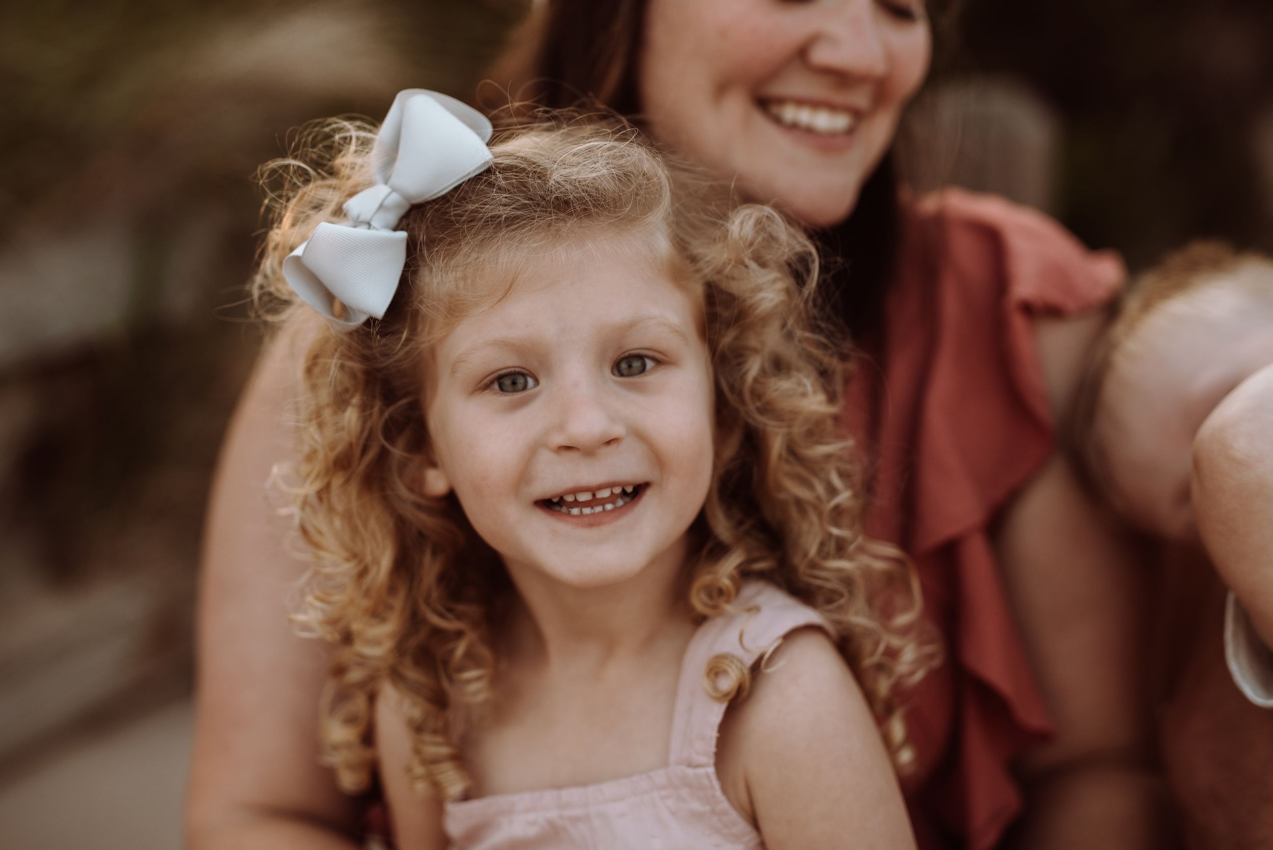 A young child with curly blonde hair and white bow smiles joyfully while wearing a light pink dress.
