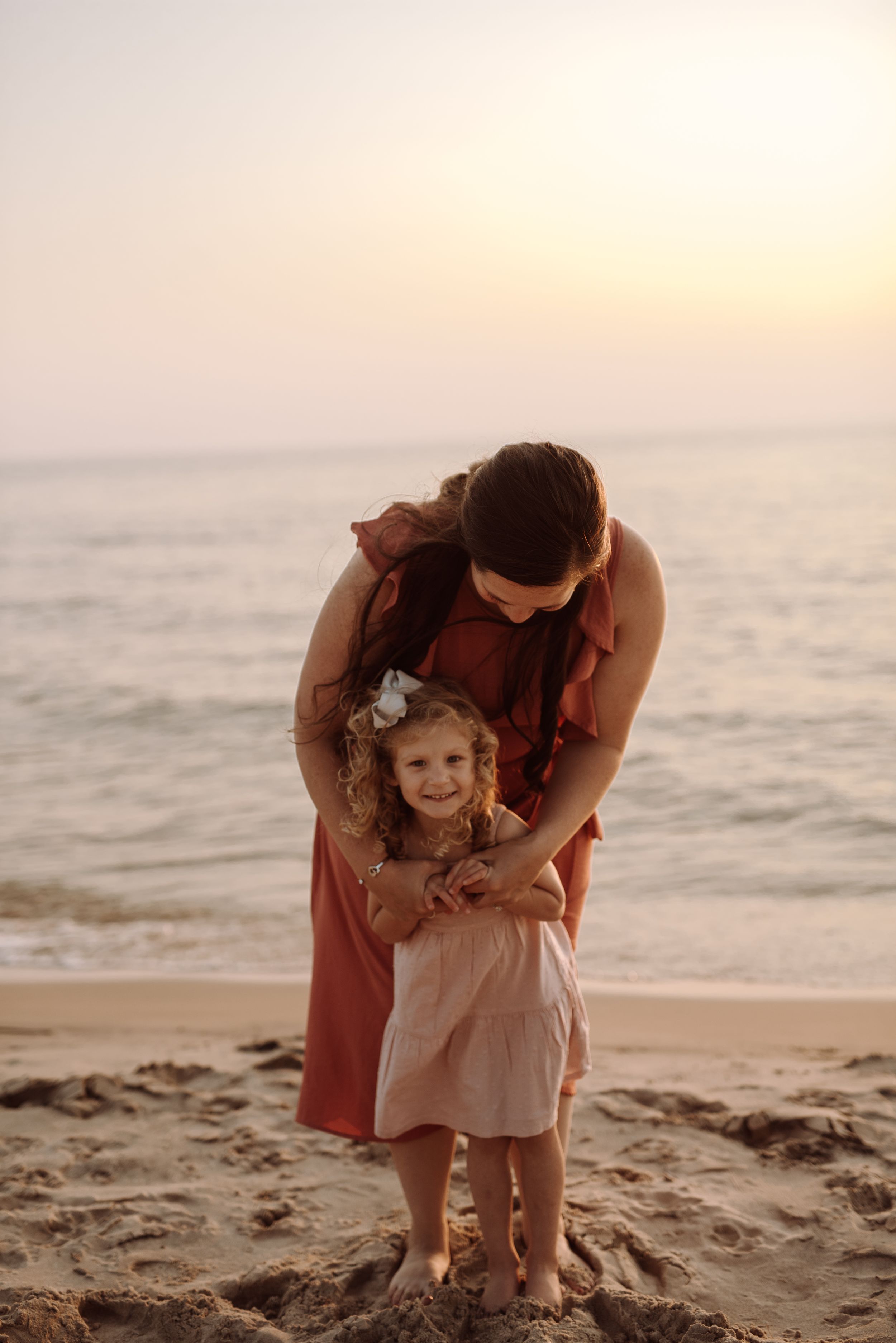 A parent and child share a tender moment on the beach at sunset while standing in the sand together.