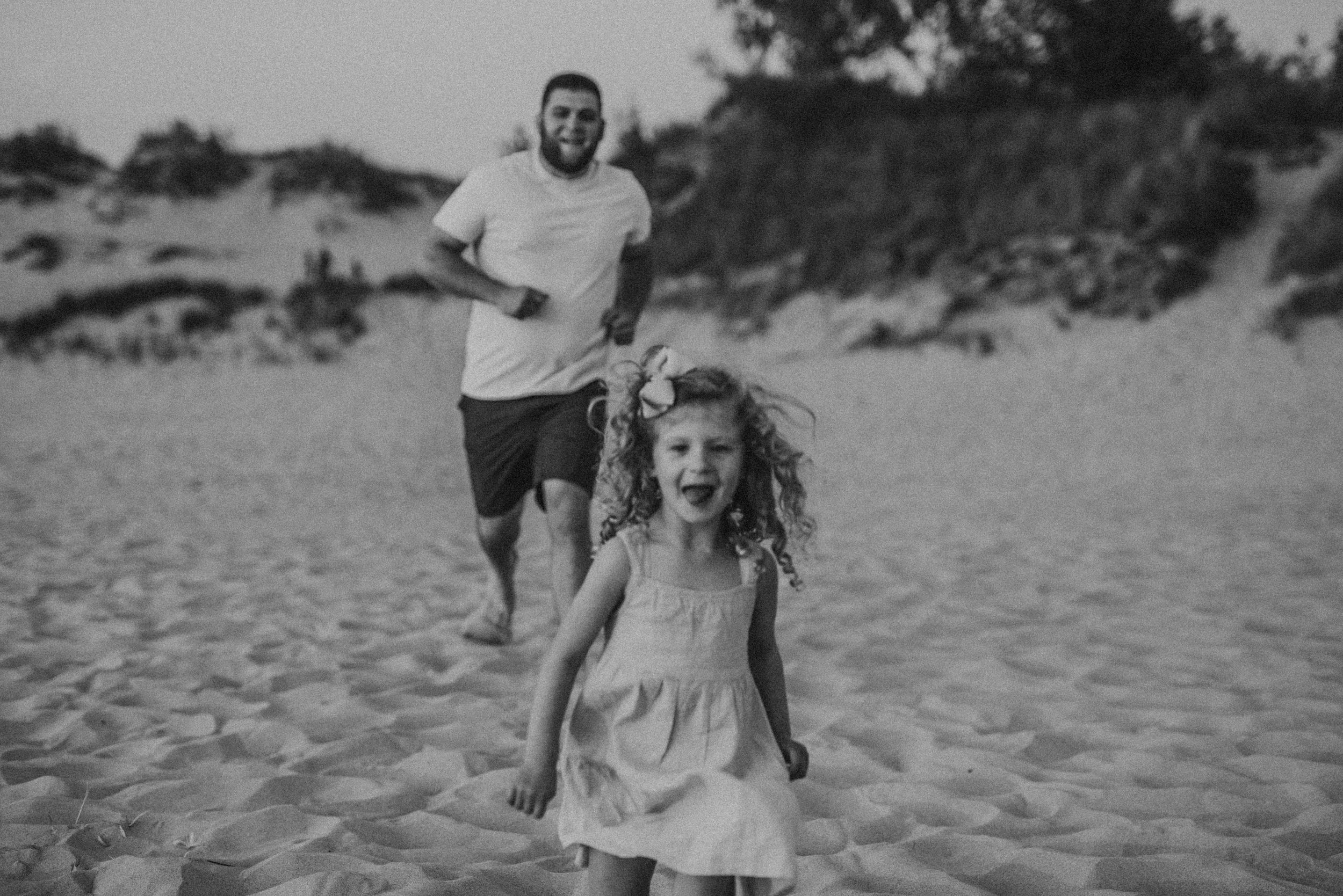 Black and white photo of a child running joyfully on a sandy beach with someone following behind.
