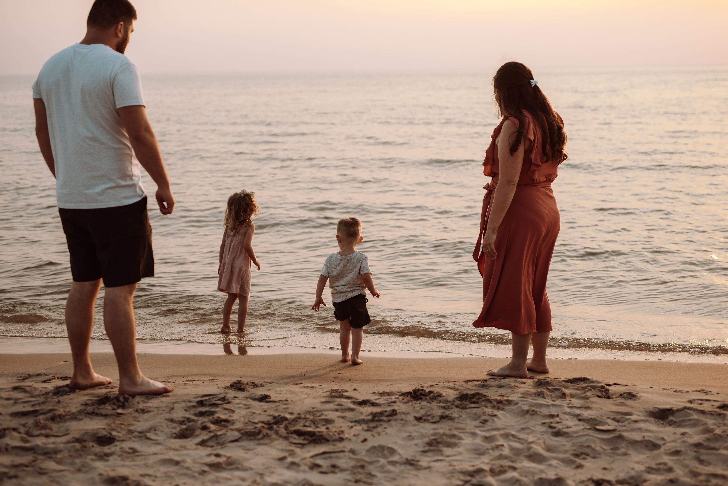 Four figures stand at waters edge during sunset on a sandy beach watching the tranquil ocean waves.