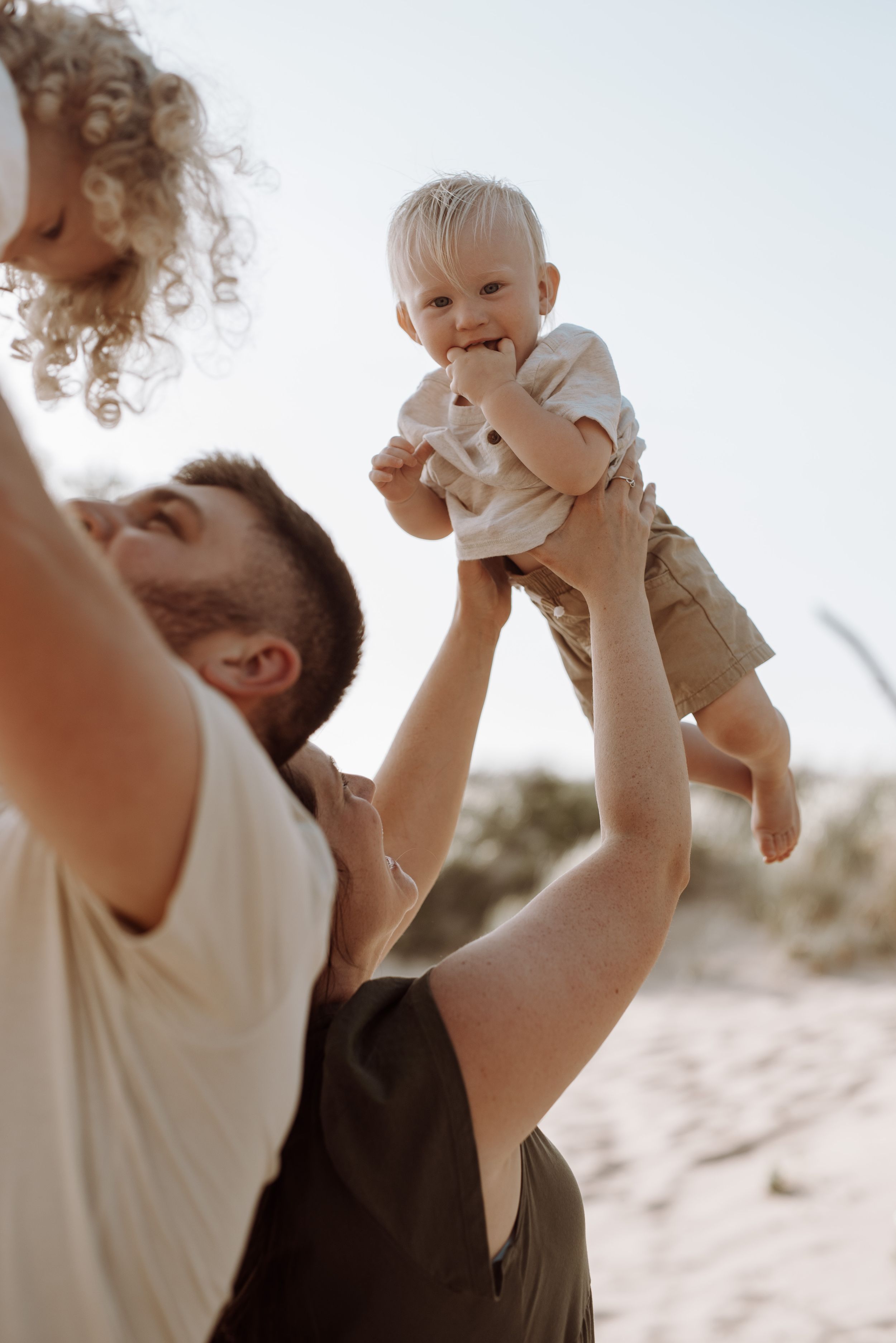 Happy child is lifted high in the air on a sunny beach as adults play together outdoors.