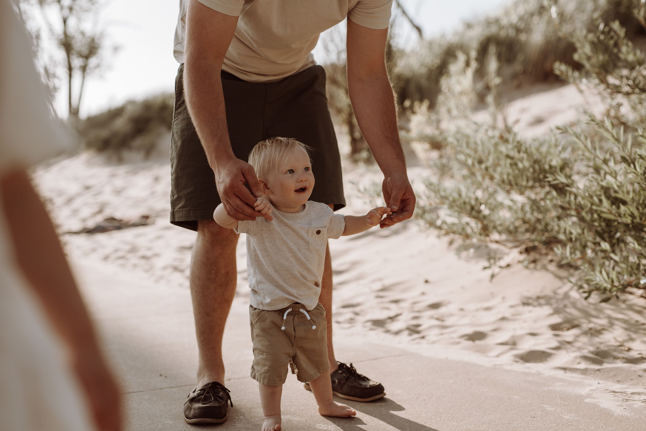 A toddler takes first steps on a sandy path while being supported by an adult wearing shorts and sneakers.