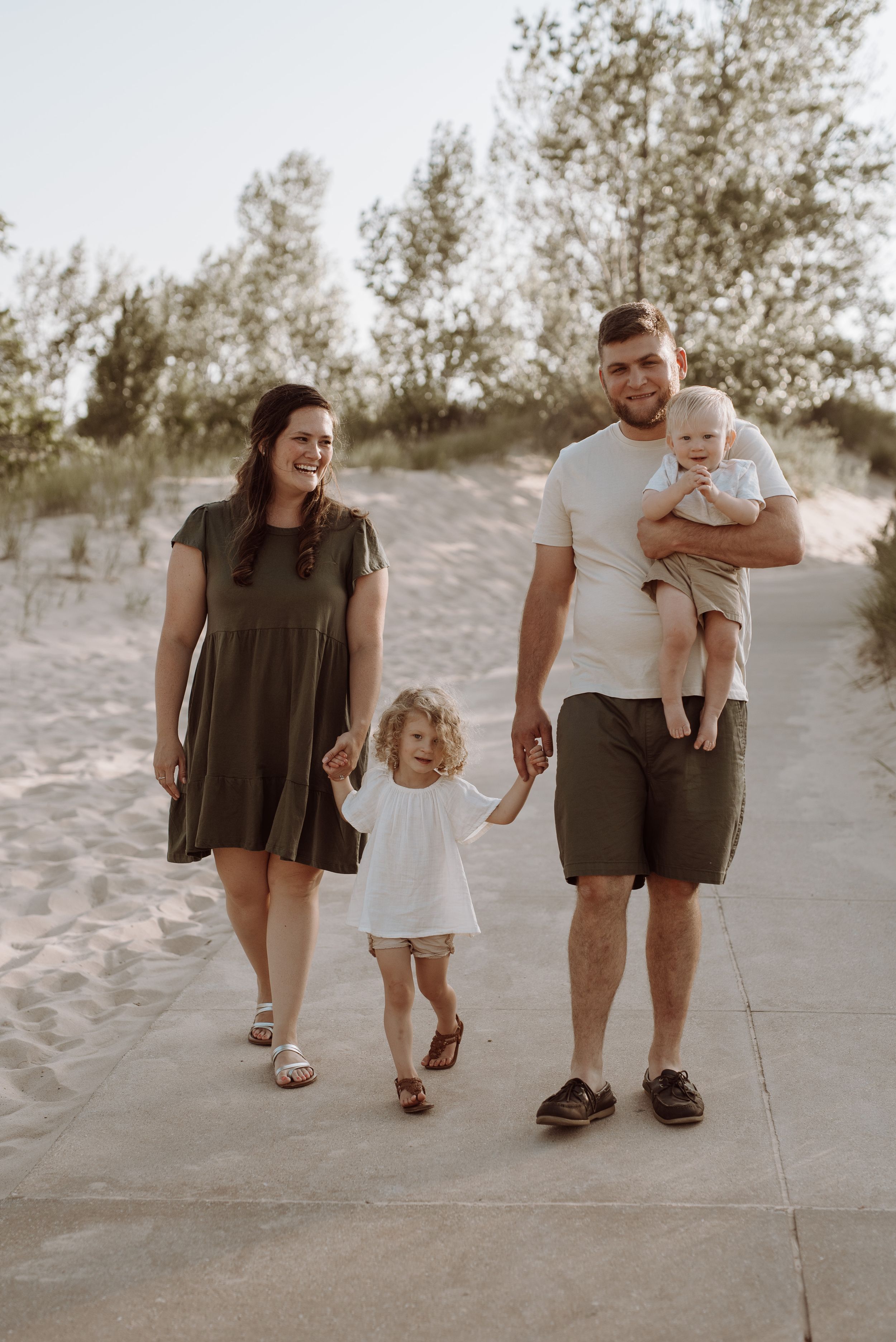 A family in neutral toned outfits walking together on sandy beach dunes during sunset in a rustic, candid photo series.