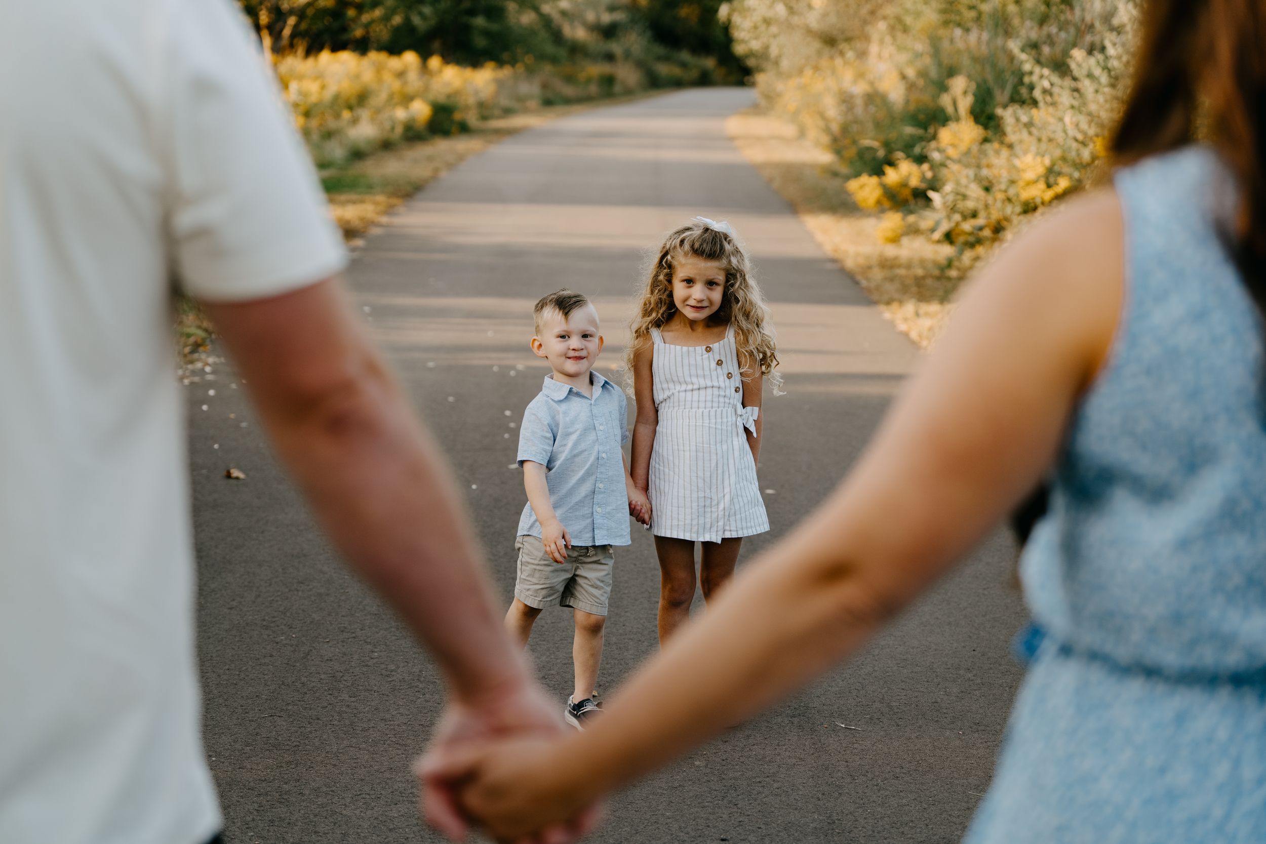 Two children walking on a tree-lined path while adults hold hands in the foreground during golden hour.
