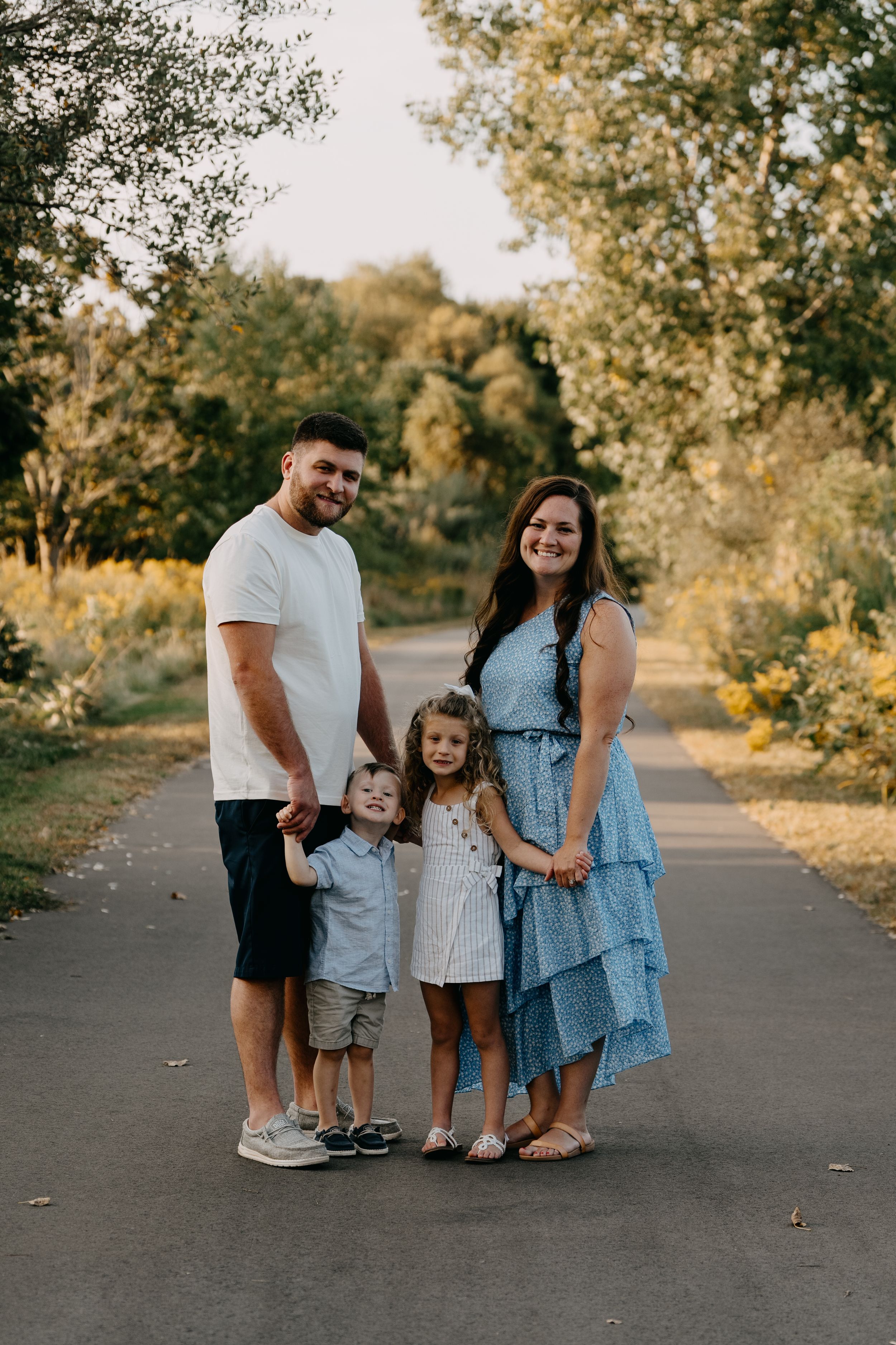 Happy family of four standing together on a paved path during golden hour surrounded by autumn trees.