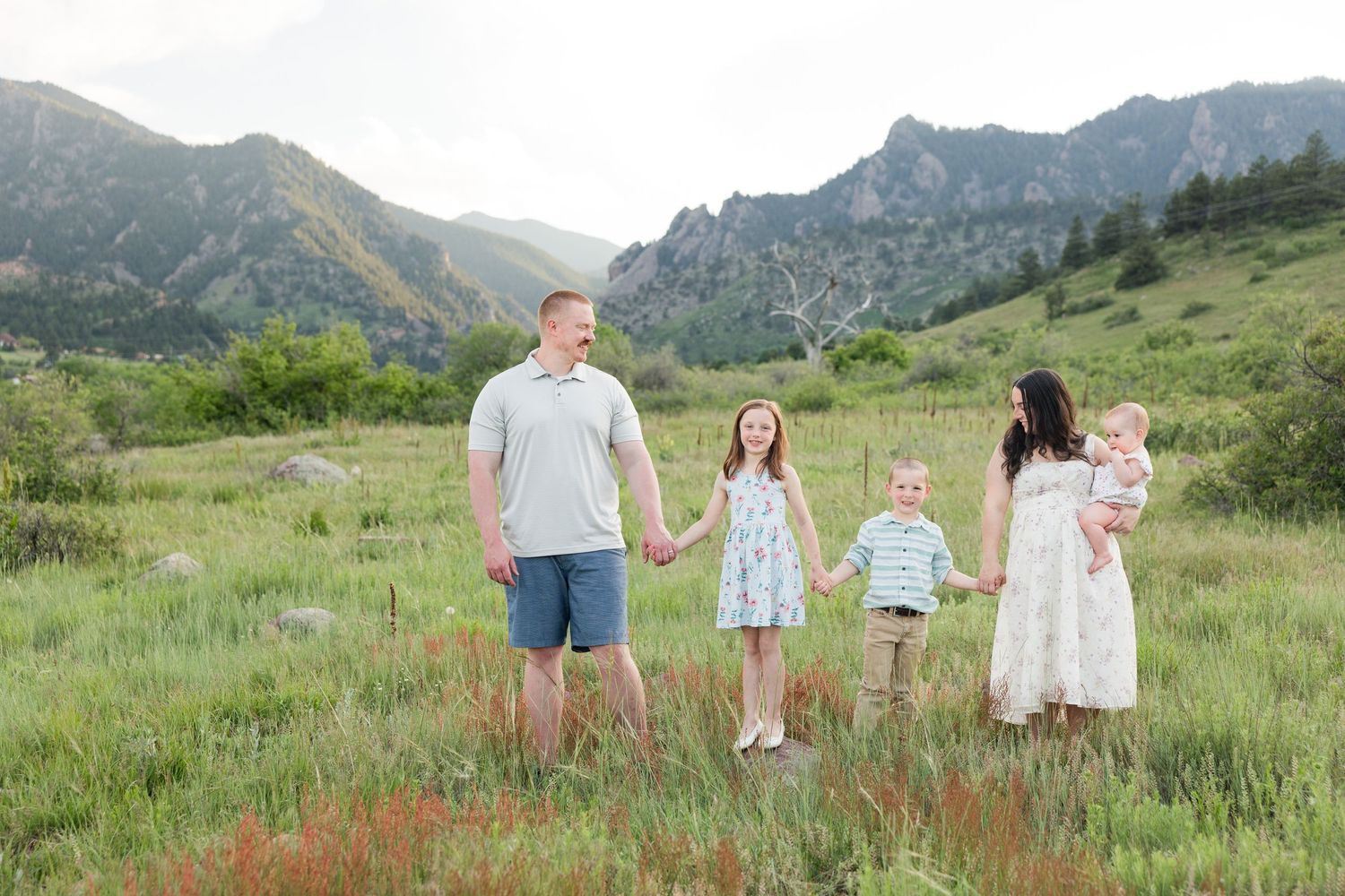 A group stands together holding hands in a scenic mountain meadow with wildflowers and rolling hills in the background.