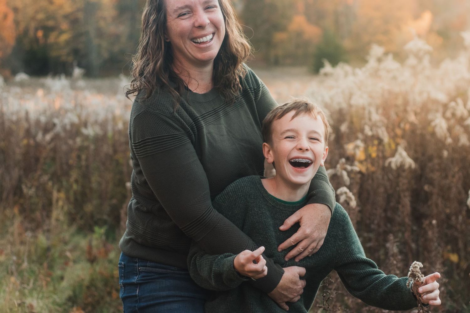 Mother and son laughing in tall grass during golden hour - lifestyle family photographer in South Lyon, Michigan.