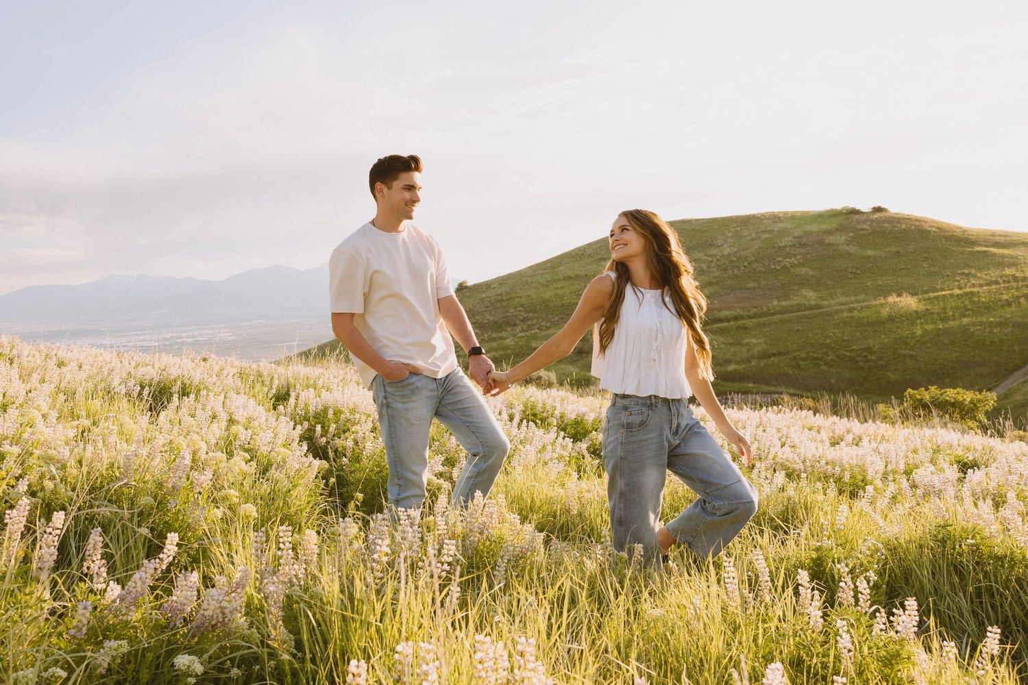 Young couple holding hands while walking through a golden sunlit meadow at sunset with rolling hills in the background.
