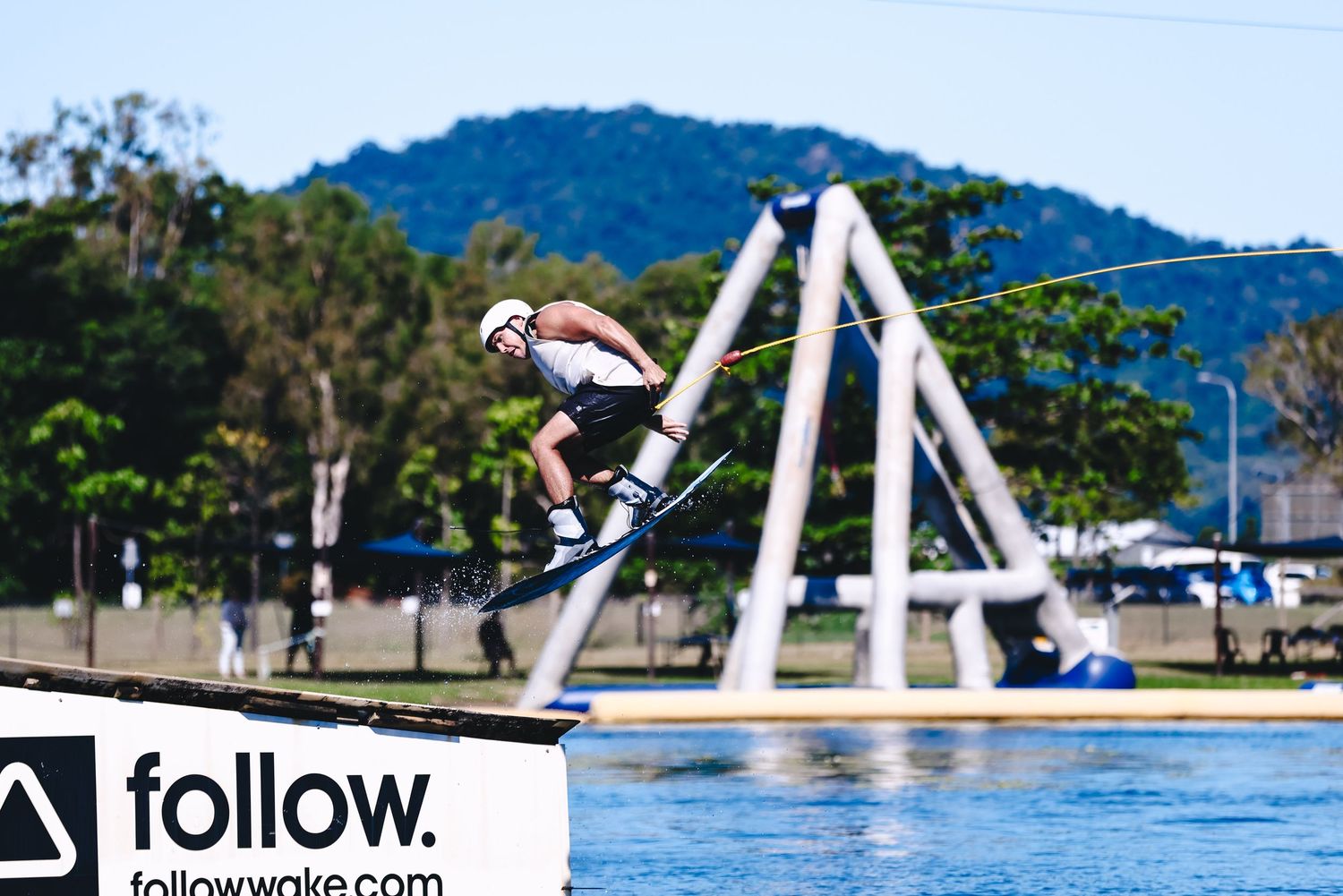 A wakeboarder performs a high-flying aerial trick over a lake with trees and power lines in the background.