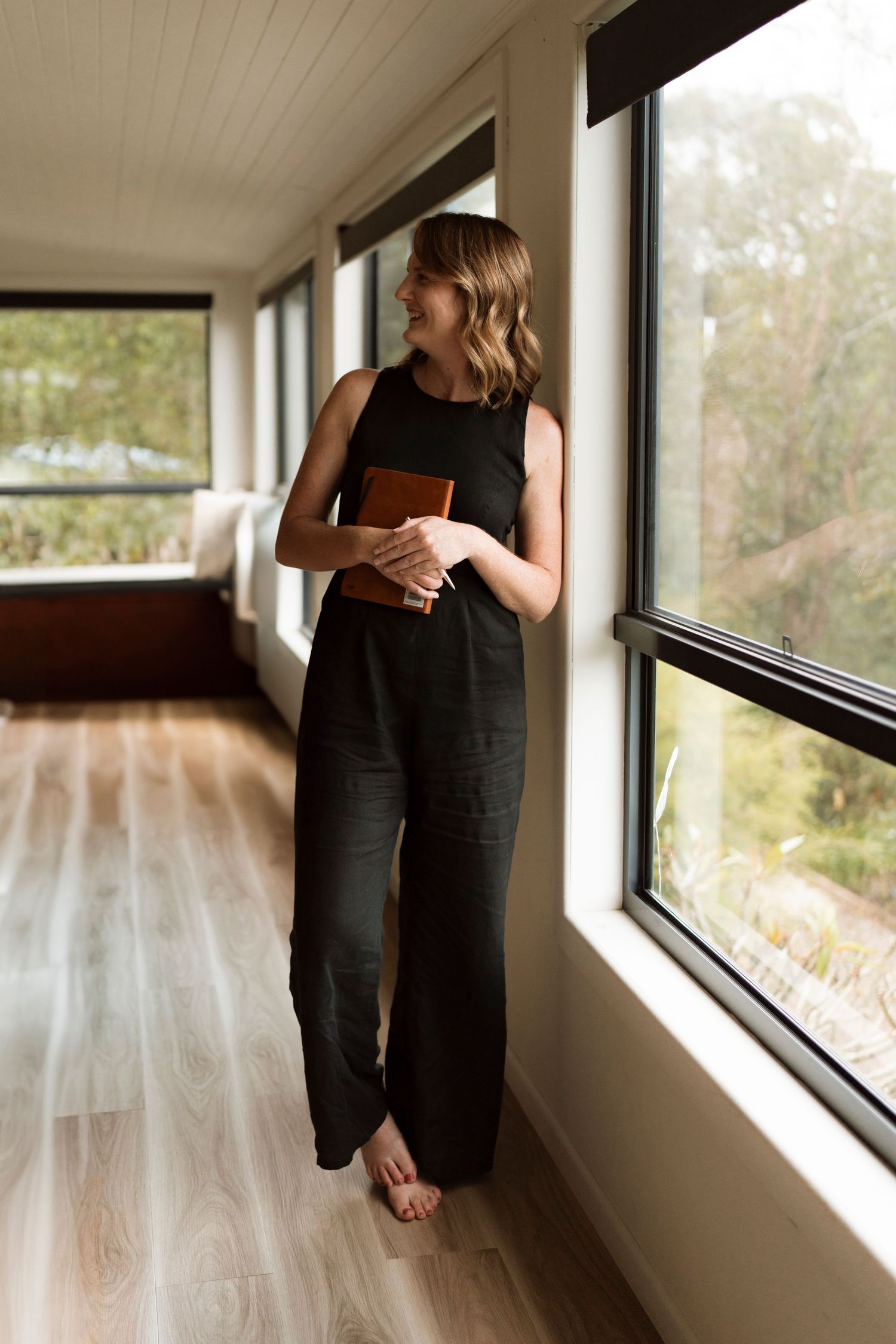 Someone in a black jumpsuit stands barefoot by large windows in a modern home with hardwood floors and natural lighting.