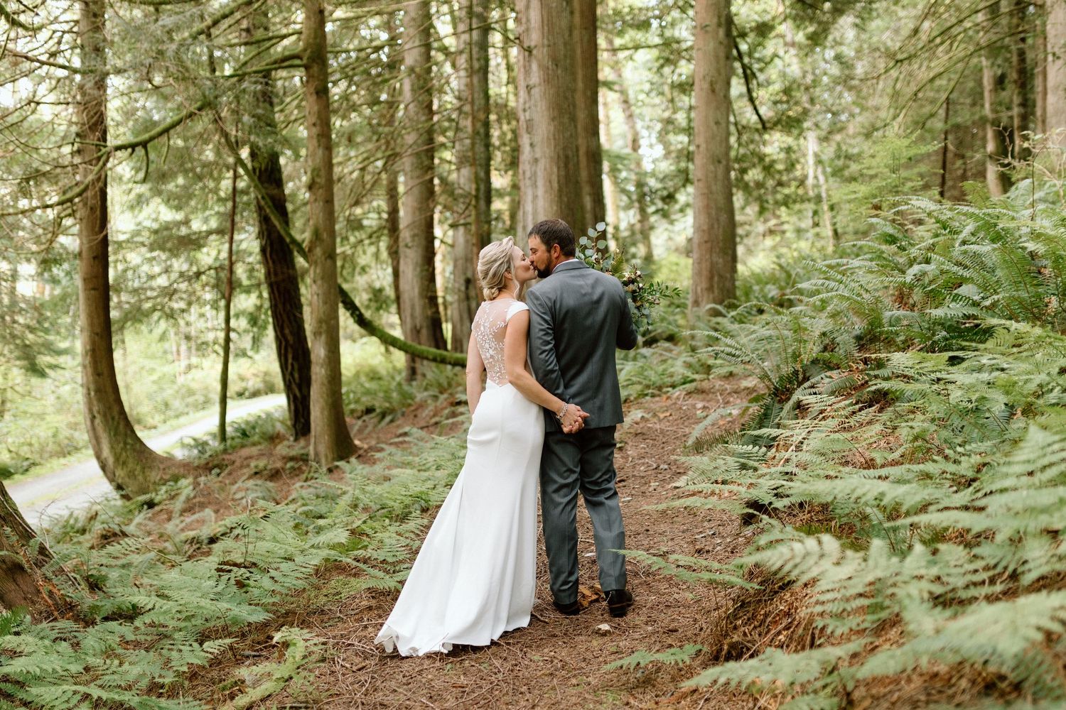In a lush forest setting with tall evergreen trees, a couple shares an intimate moment on a trail surrounded by ferns.