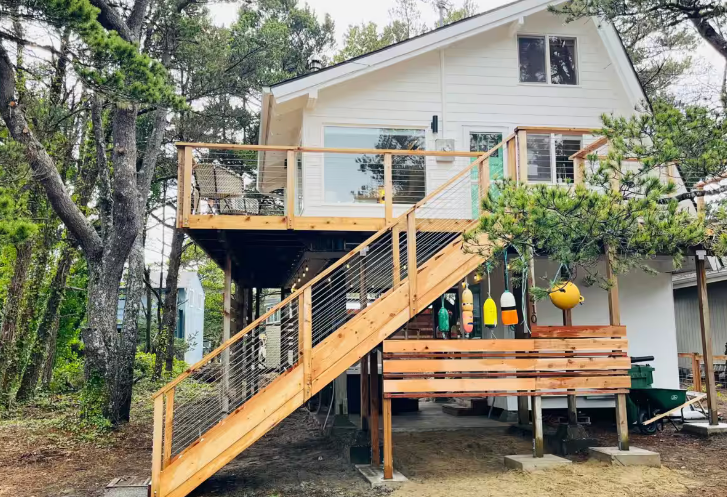 White two-story beach house with wooden deck and stairs featuring yellow cushioned seating and forest backdrop.