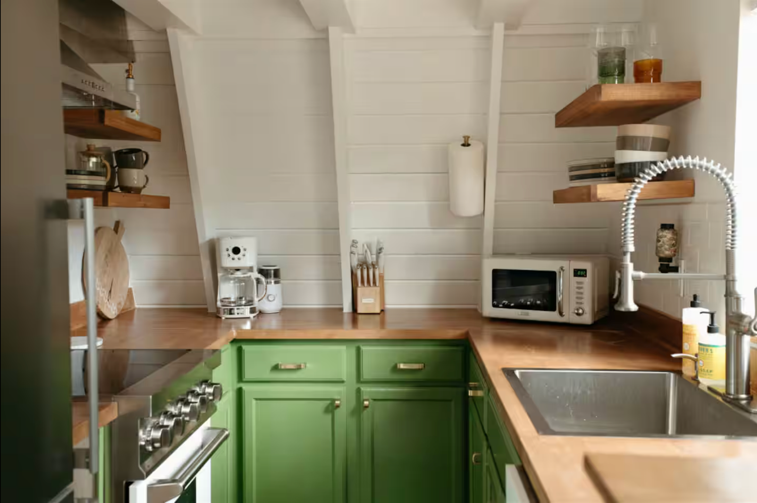 Modern kitchen with green lower cabinets, butcher block countertops, white subway tile walls and floating wood shelves.