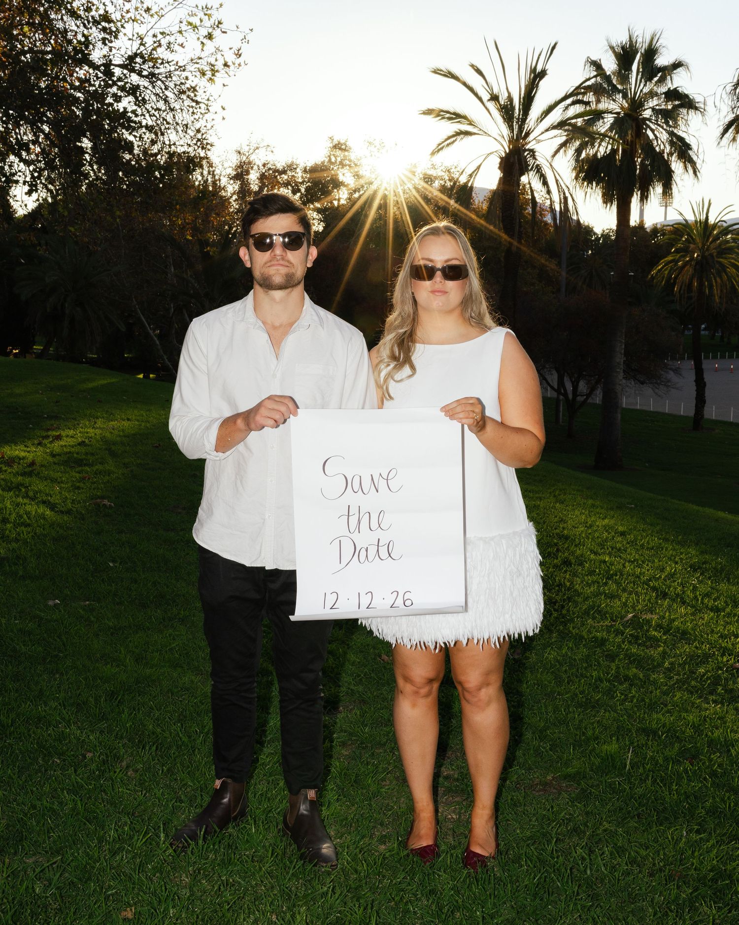 Couple holds a 'Save the Date' sign in front of palm trees during sunset in a lush garden setting.