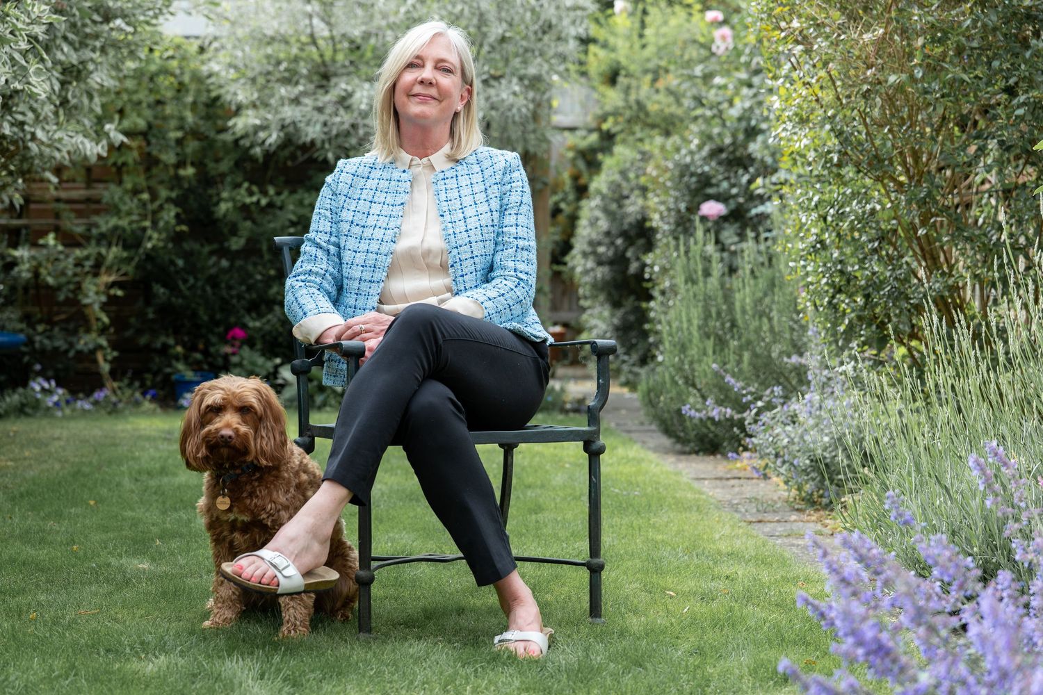 A person sits in a garden chair with a brown Cockapoo dog beside lavender flowers and lush greenery.