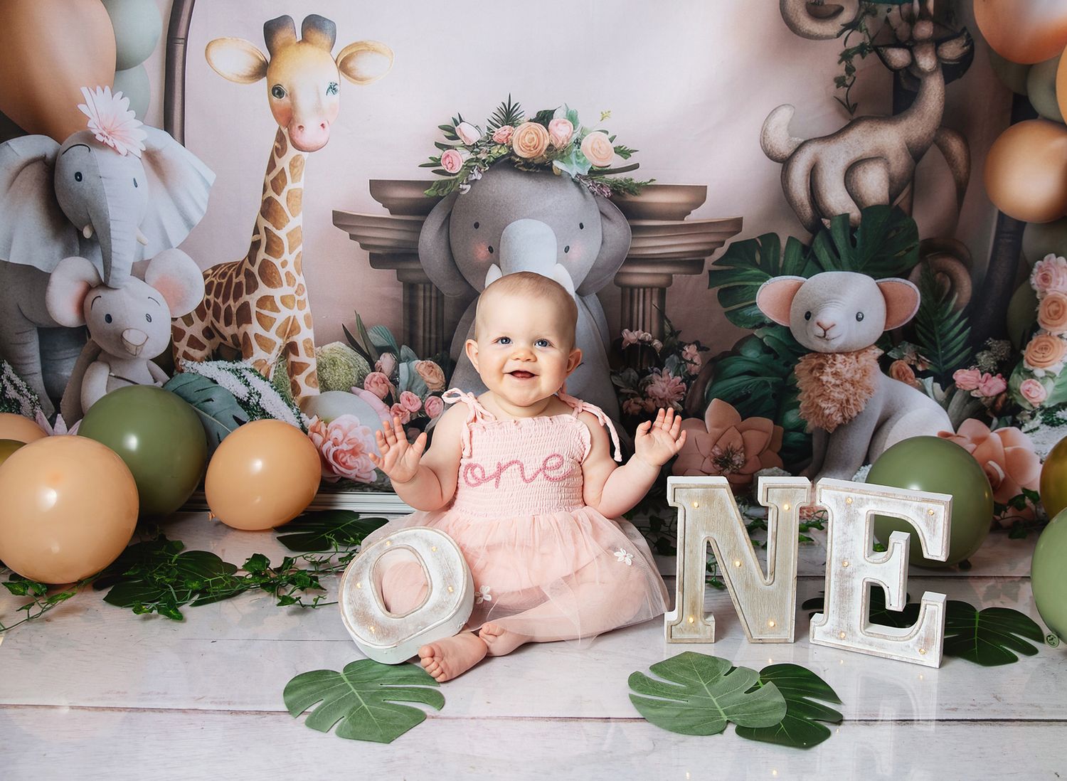 Little one sitting among decorative stuffed animals, balloons and 'ONE' letter props in a whimsical birthday photo setup in Bexley