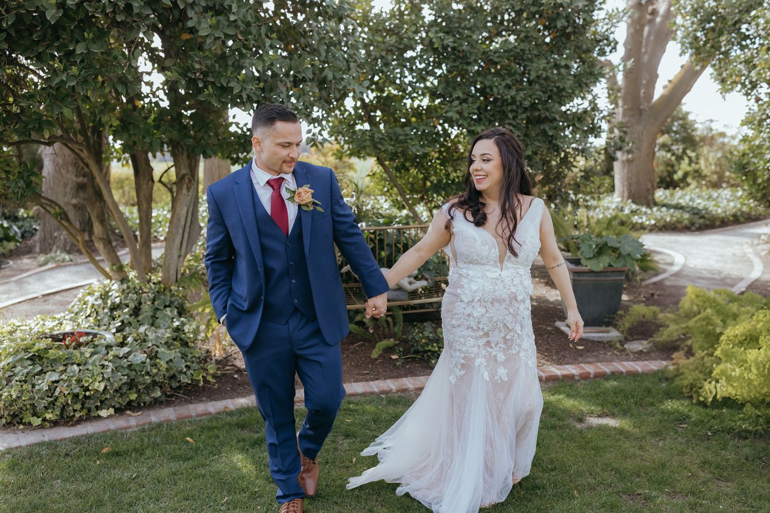 couple in wedding attire walking hand in hand through a garden surrounded by trees and greenery