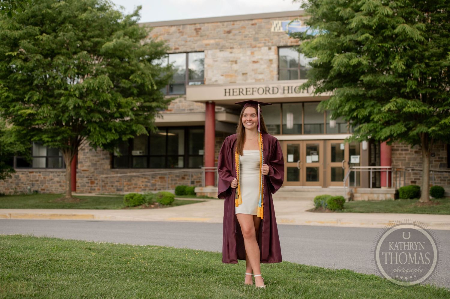 Graduate poses in maroon cap and gown outside Hereford High School entrance on graduation day.