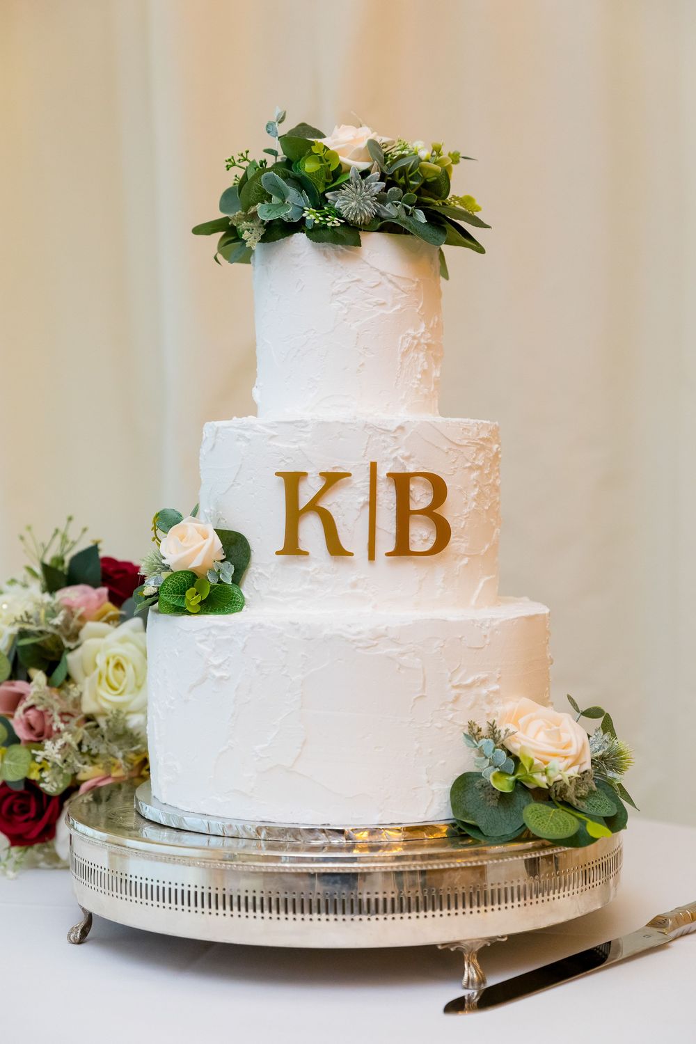 Elegant three-tier white wedding cake with floral touches and gold initials K and B at Audley’s Wood Hotel.