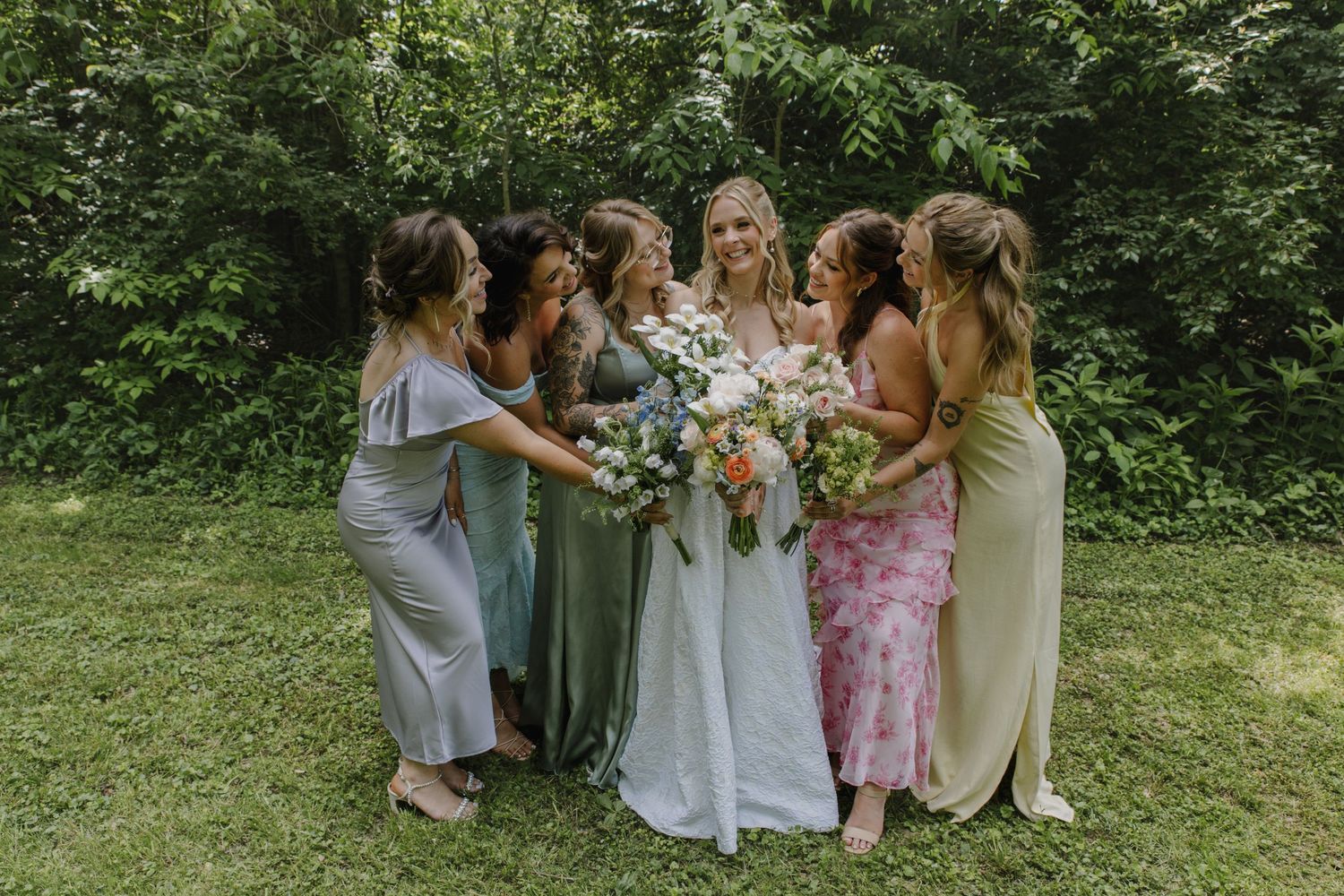 A bridal party in pastel dresses gathers for a joyful photo in a garden setting.