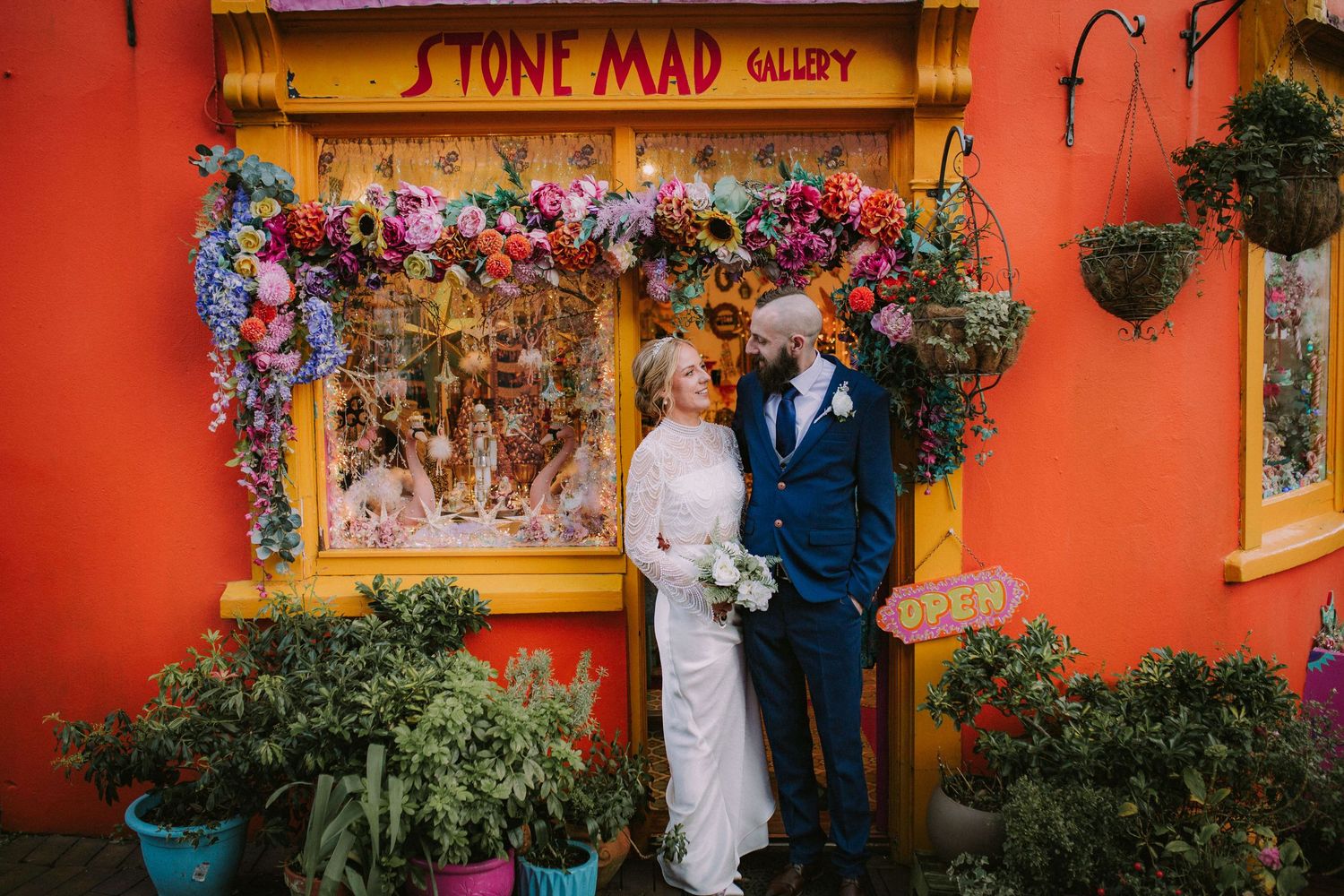 A couple stands outside a vibrant orange Stone Mad Gallery storefront decorated with colorful floral arrangements.