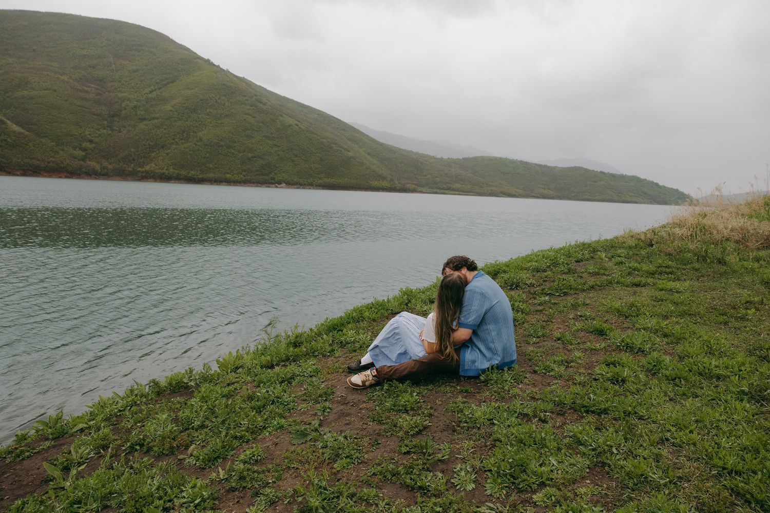 Couple sitting together on a grassy lakeside bank with misty green hills in the background.